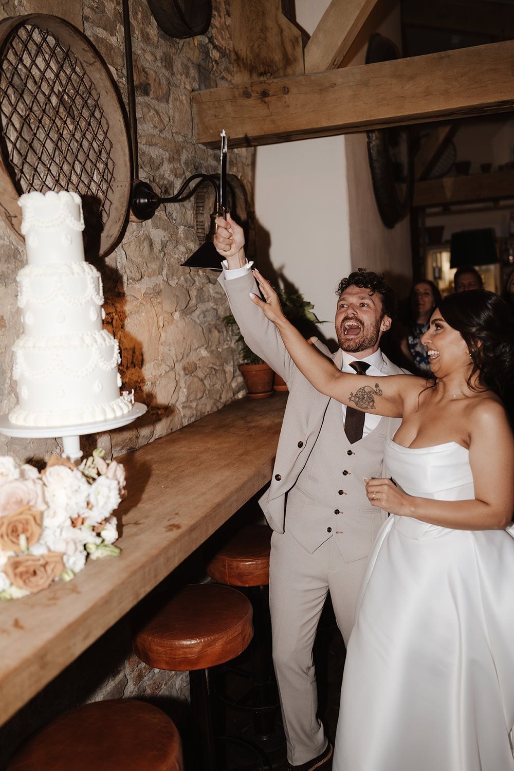 A joyful couple cuts their elegant white wedding cake together in a rustic stone-walled venue.