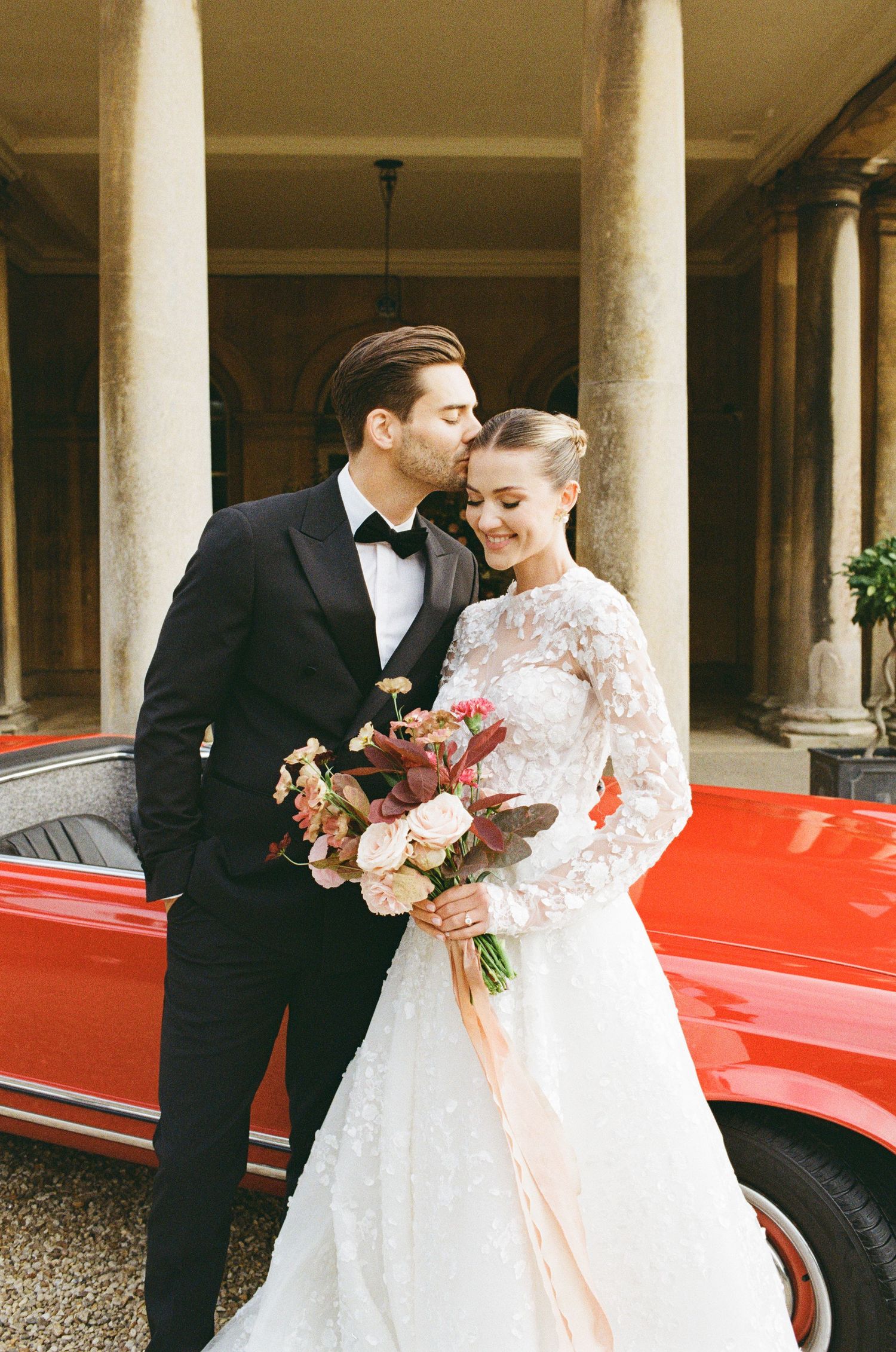 A couple share an intimate moment beside a red vintage car, with elegant white columns framing the romantic scene.