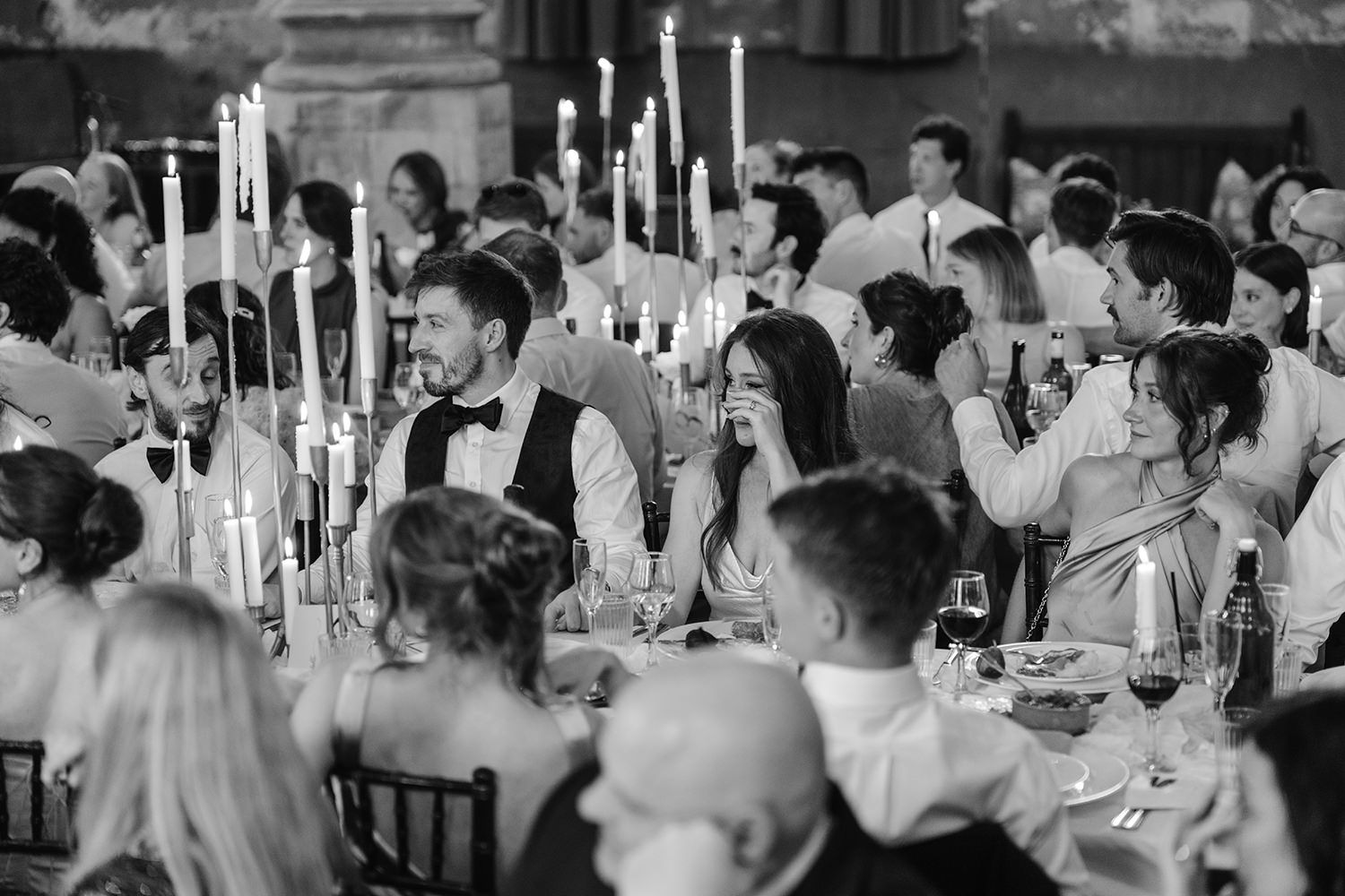 Wedding guests seated at elegant tables with white candles celebrate during an indoor reception in black and white.