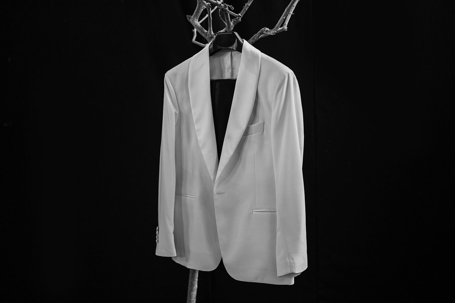 White wedding blazer hung up on a tree against a solid black backdrop.