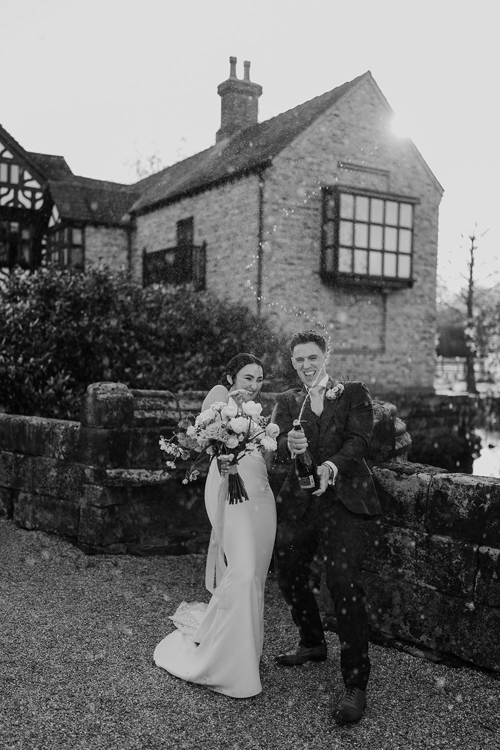 Bride and Groom standing laughing with a bottle of champagne spraying with Holford Hall behind.