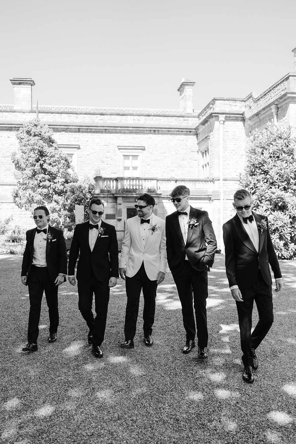 A sequence of groomsmen in black tuxedos walking and posing on a grassy lawn outside an elegant building.