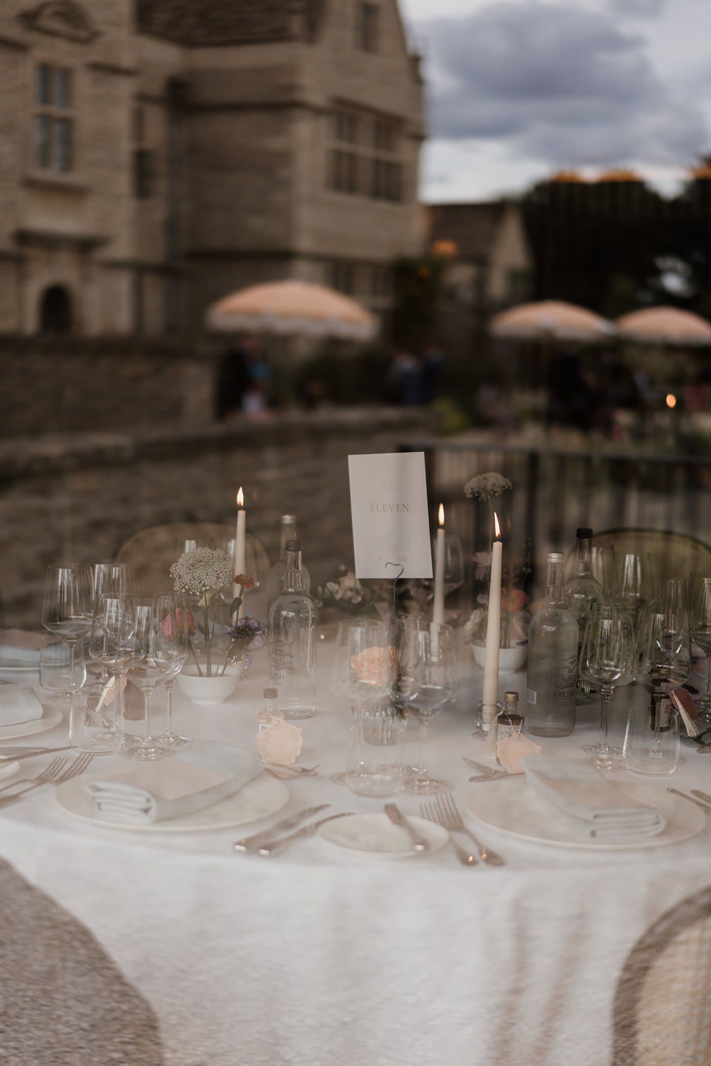 Elegant wedding table setting with white linens, glassware, and candlesticks against a castle backdrop at dusk.