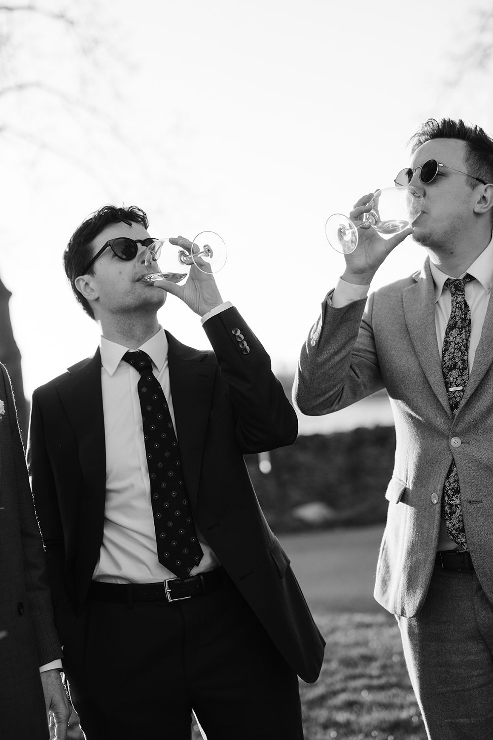 Two stylish businessmen in suits and sunglasses drinking beverages outdoors in black and white photography.