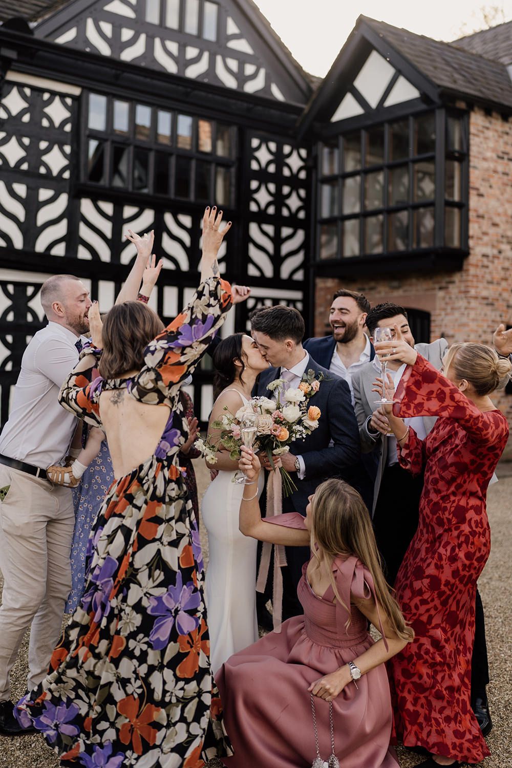 Bride and groom kissing as friends dressed in bright wedding outfits laugh and dance with Holford Hall behind them.