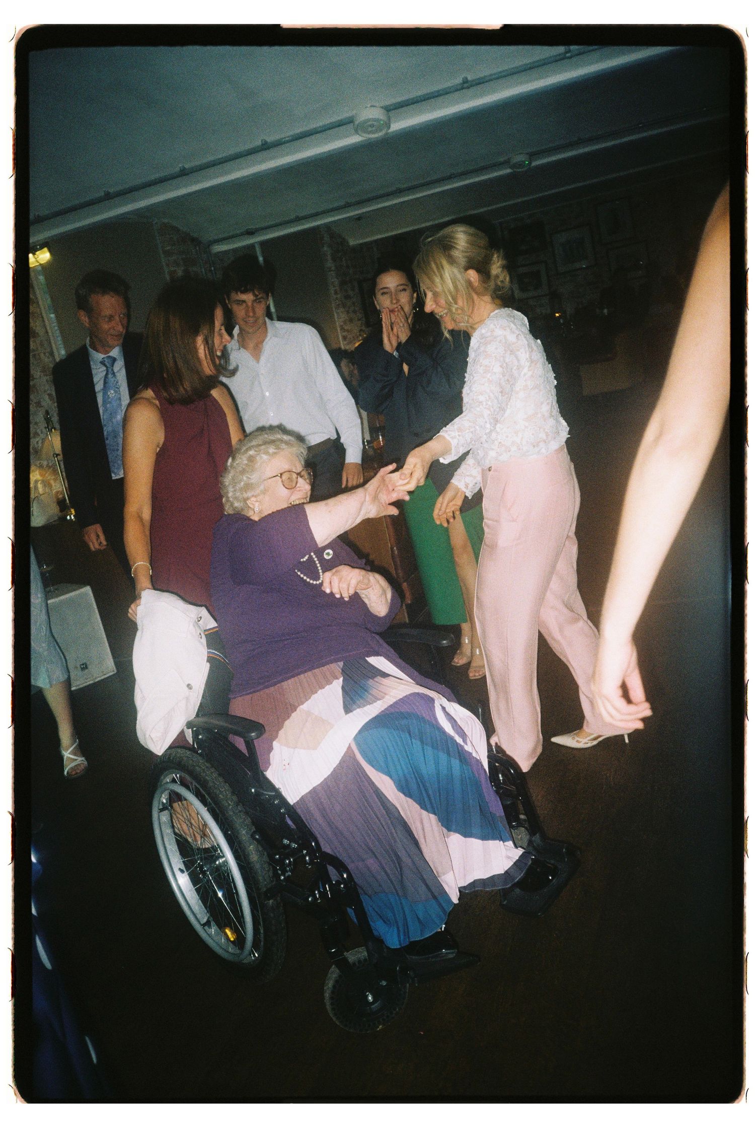Elderly woman in wheelchair holding hands with young woman in pink at a lively indoor party gathering.