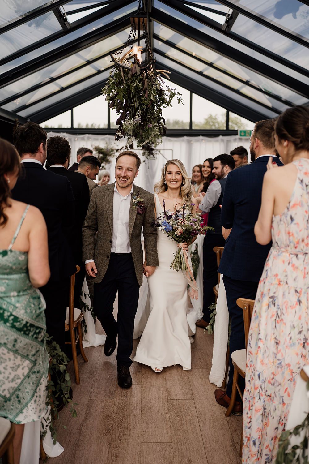 Bride and groom holding hands walking out of their ceremony with guests stood clapping at either side.