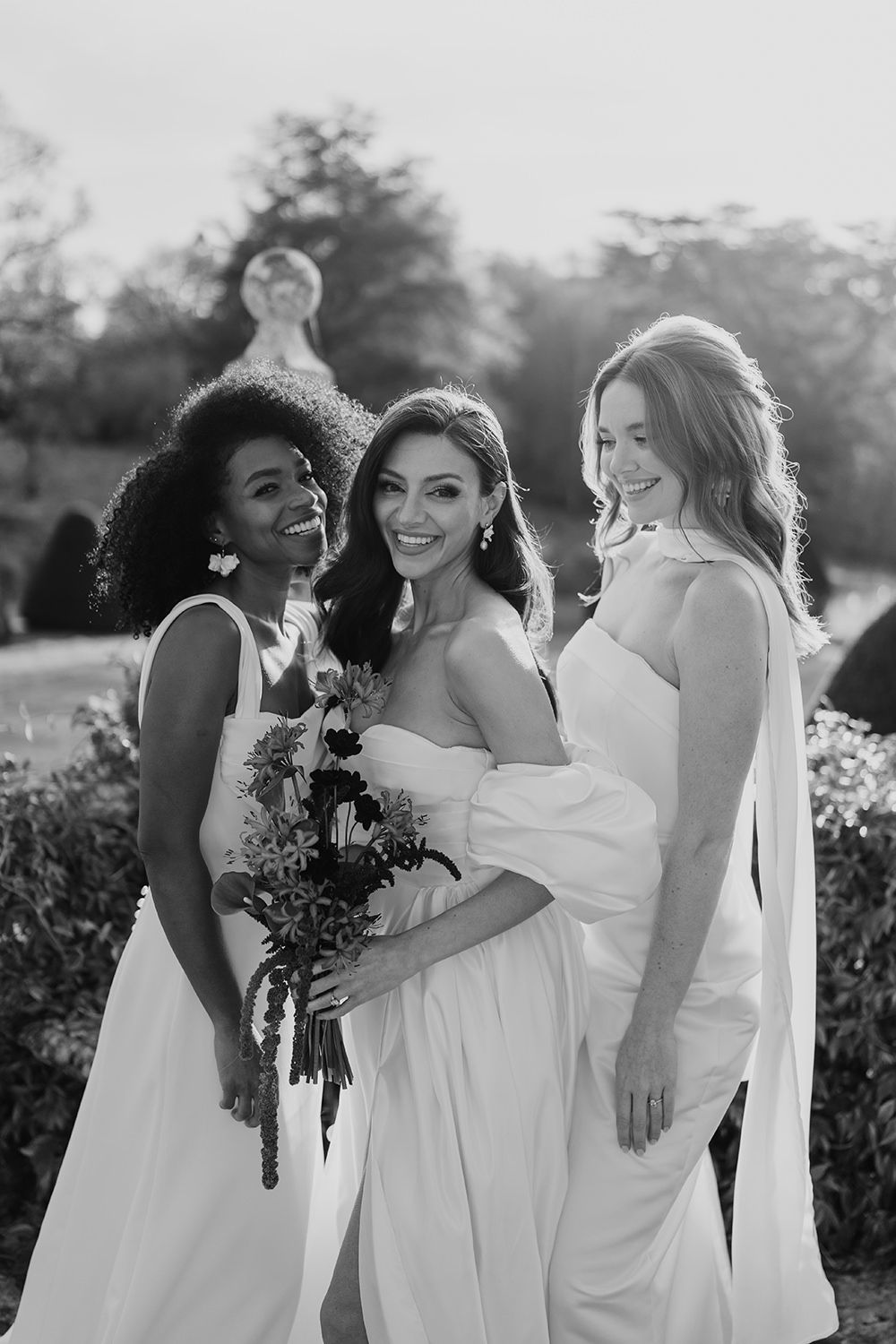 Black and white wedding photo of bridesmaids in elegant white dresses posing outdoors with flowers and joyful expressions.