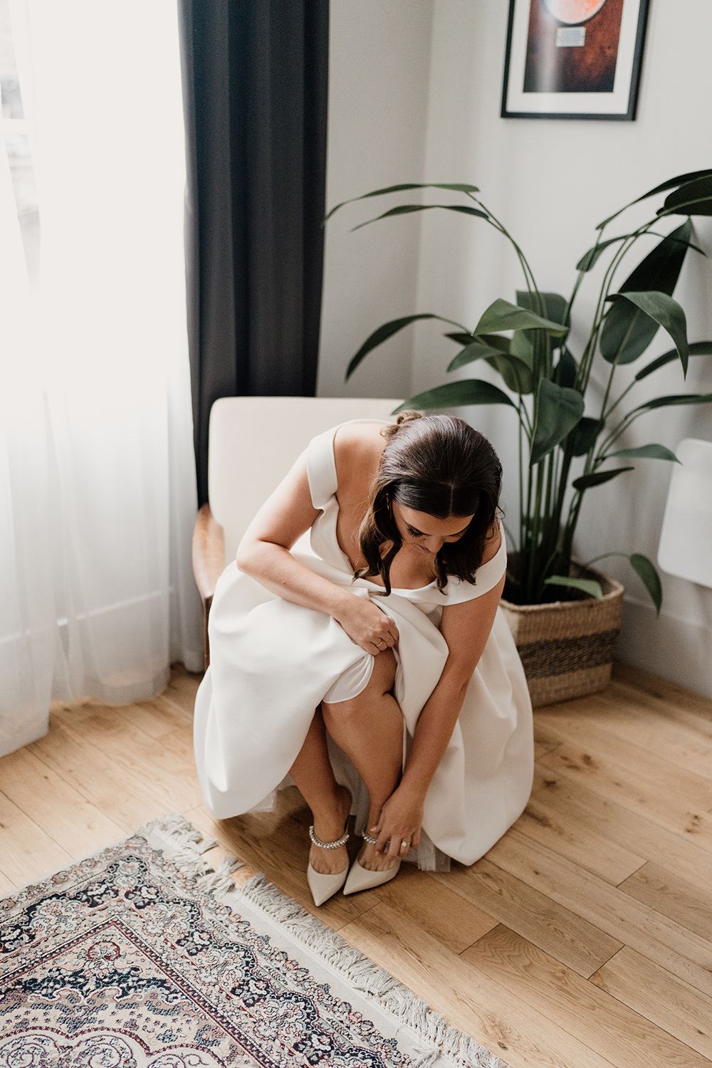 Person in white dress sitting in modern living room with houseplants adjusts footwear on hardwood floor by patterned rug.