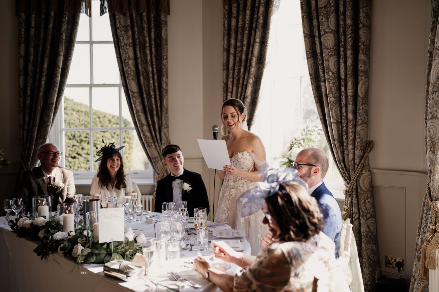 Bride making a speech on her wedding day with family around her and windows behind.