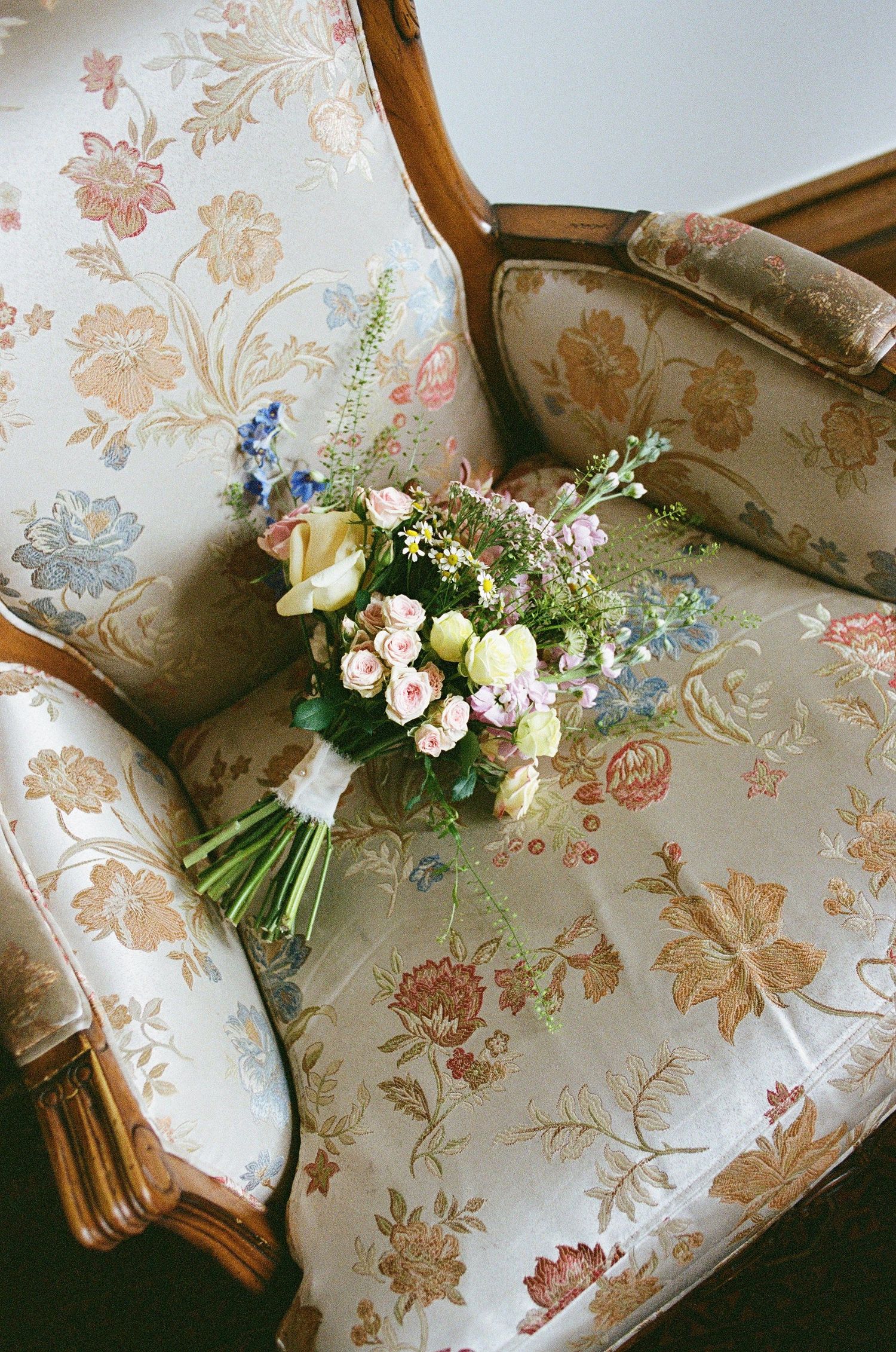 Romantic bridal bouquet of pink roses, tulips, and wildflowers resting on a vintage floral upholstered armchair.