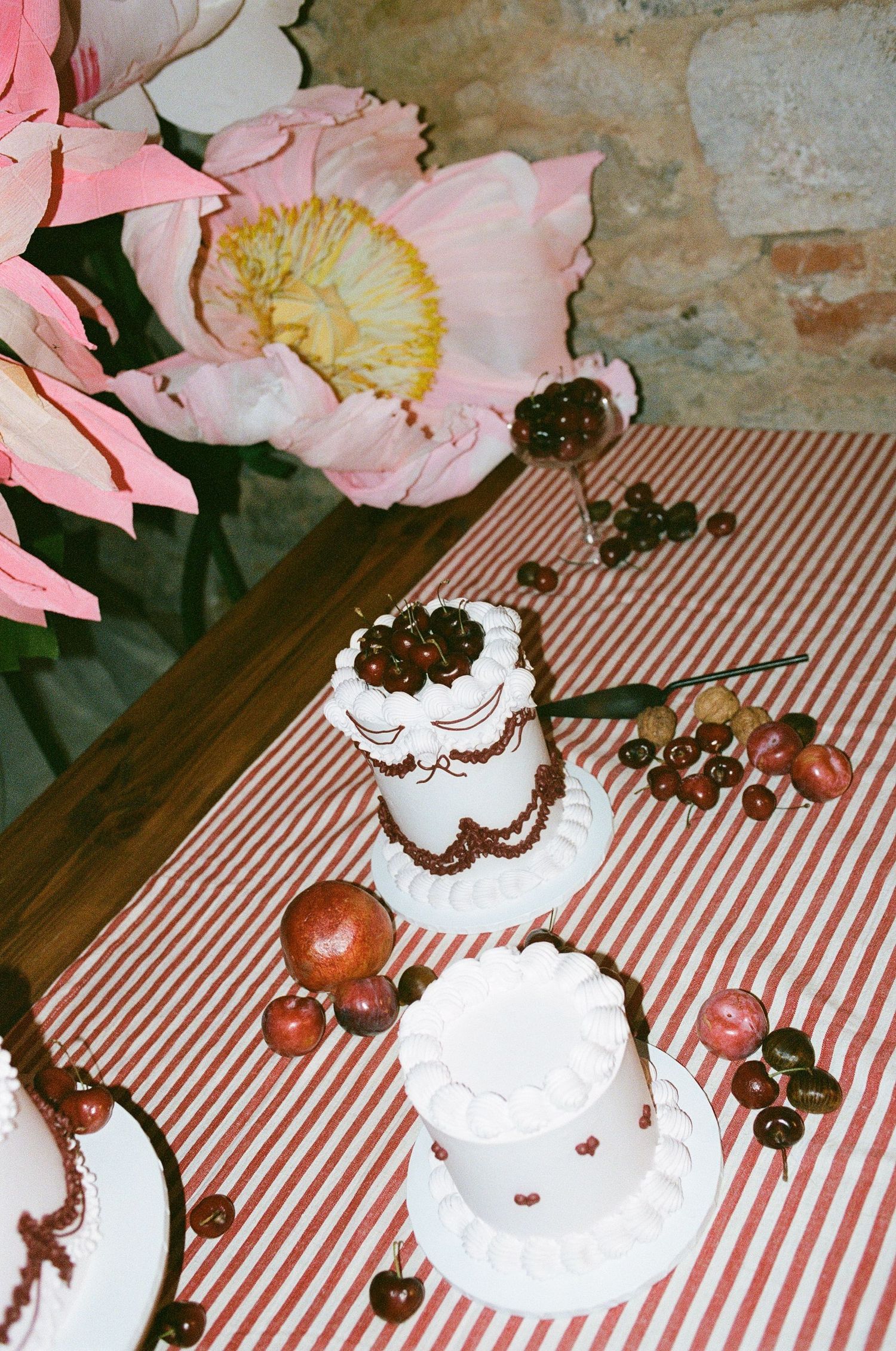 Miniature white wedding cakes decorated with cherries on a red striped tablecloth with large pink florals.