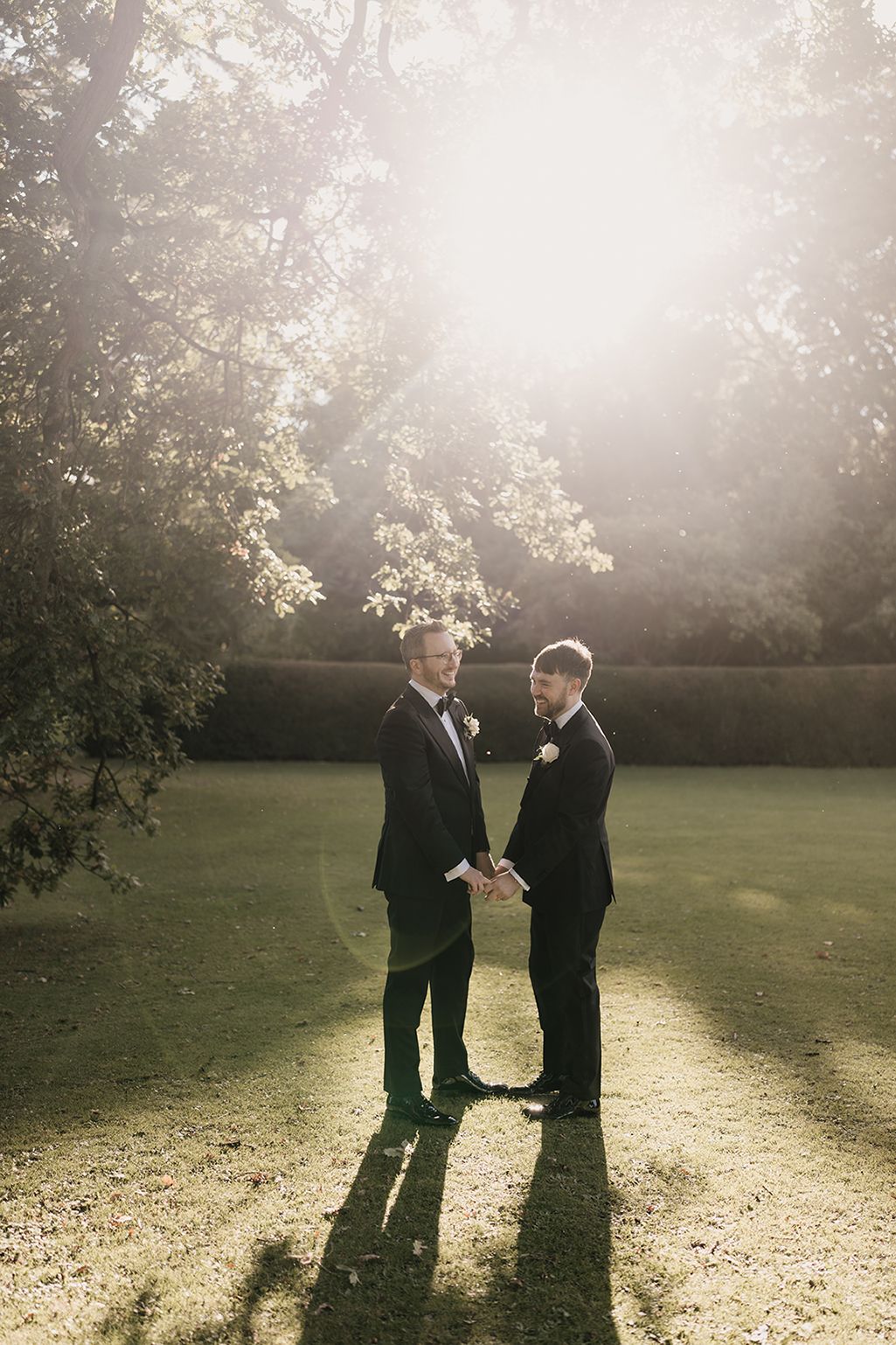 Two grooms in black tuxedos hold hands in a sunlit garden during their outdoor wedding ceremony.