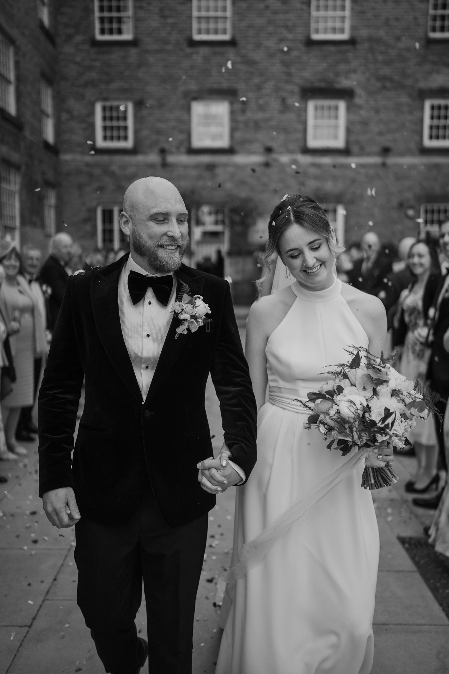 Happy newlyweds walking together while guests throw confetti outside brick building.
