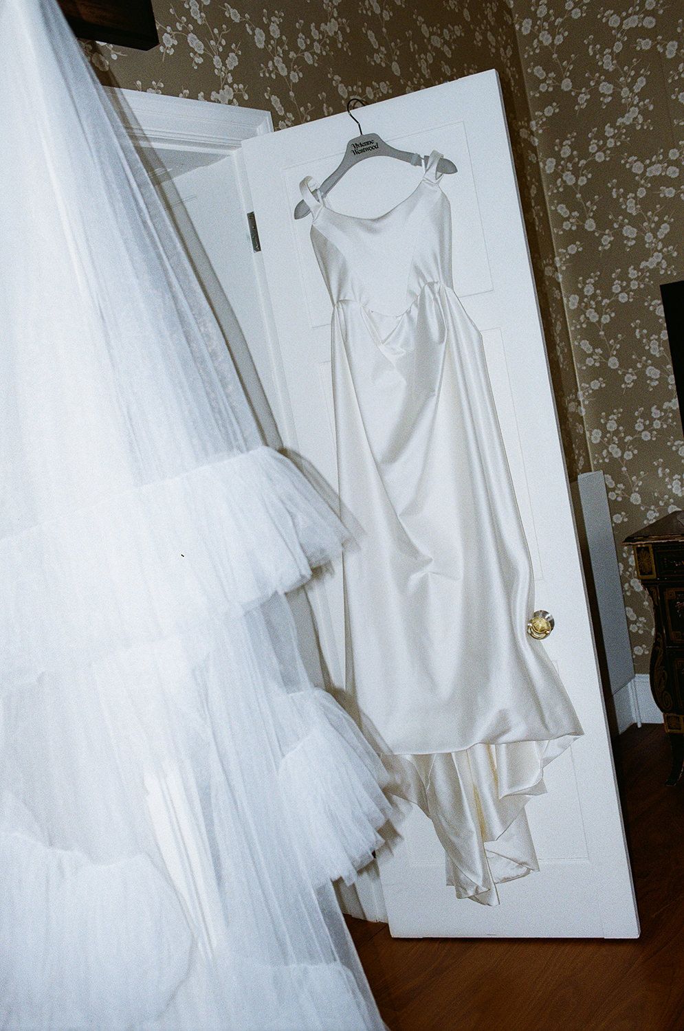 A white wedding dress hangs on a door next to a tulle veil in a vintage-style photograph.