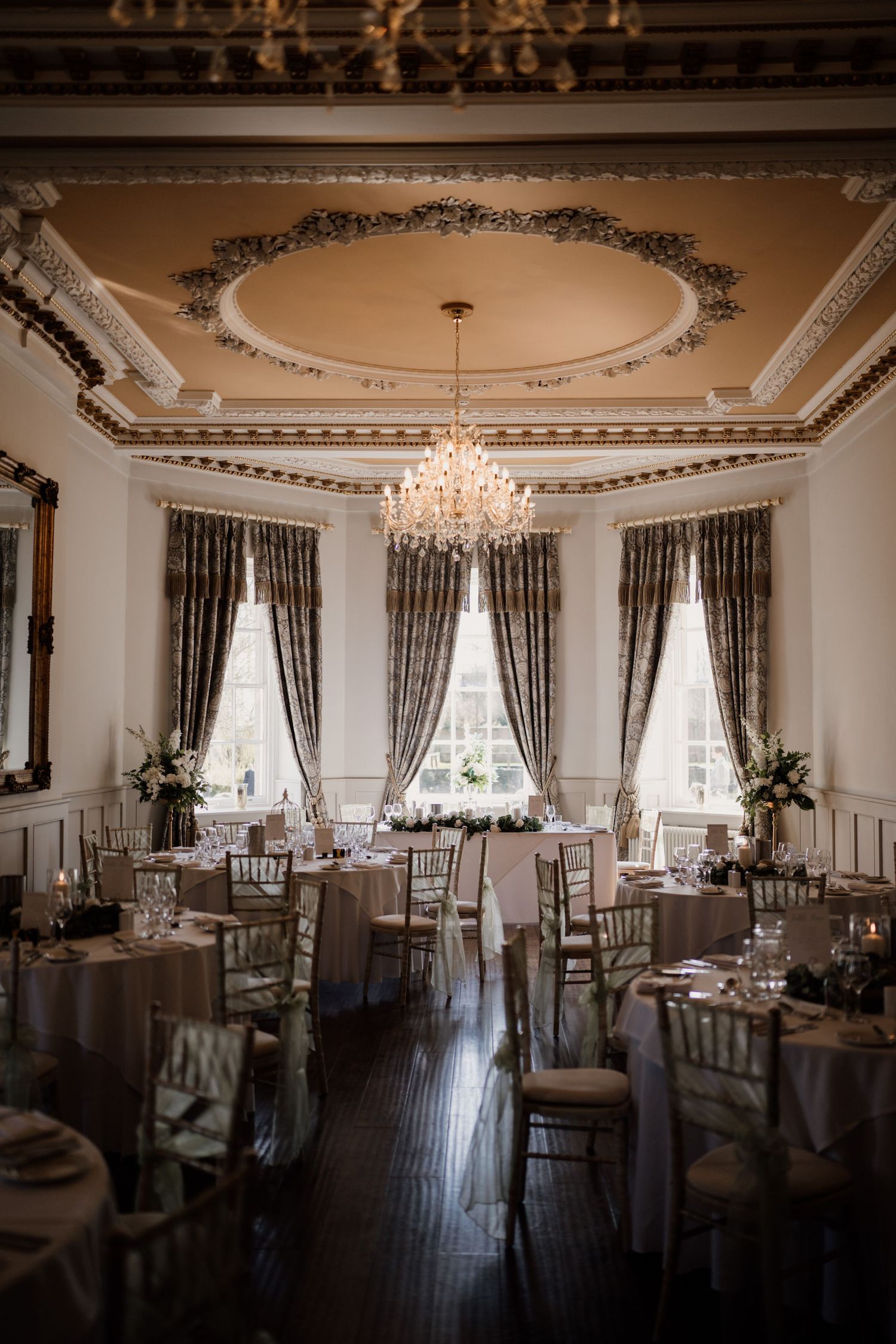 Room shot of Bourton Hall ready for wedding speeches with round tables styled and large windows dressed.