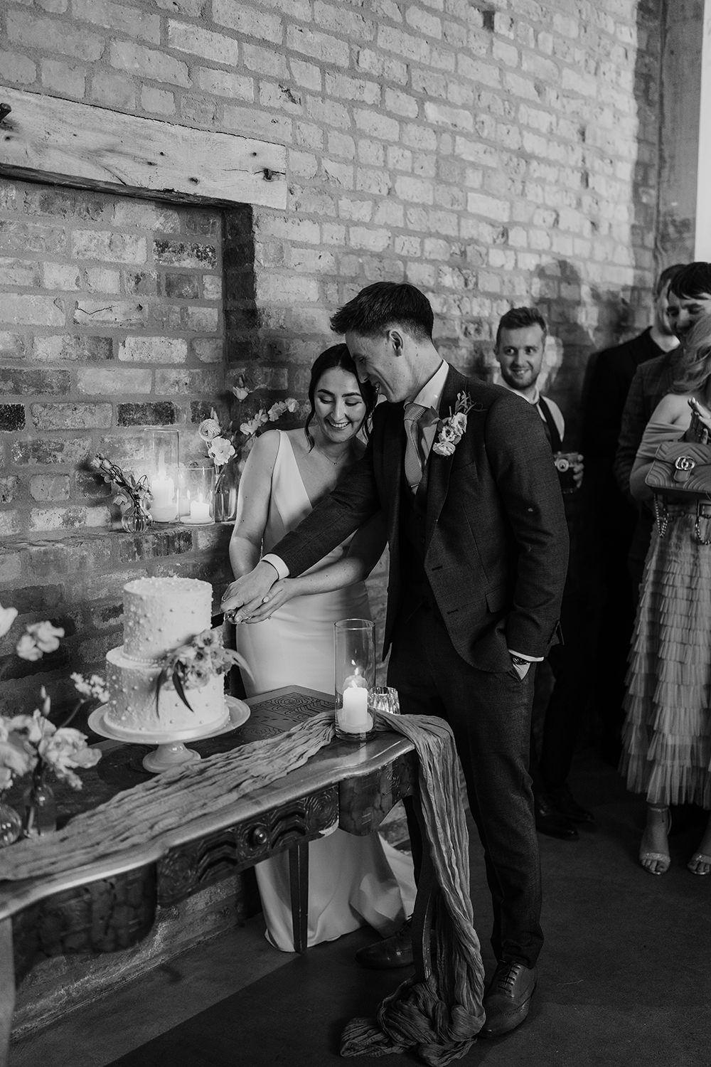 Bride and groom stood cutting their wedding cake with guests watching, the cake is on a table with flowers and candles.