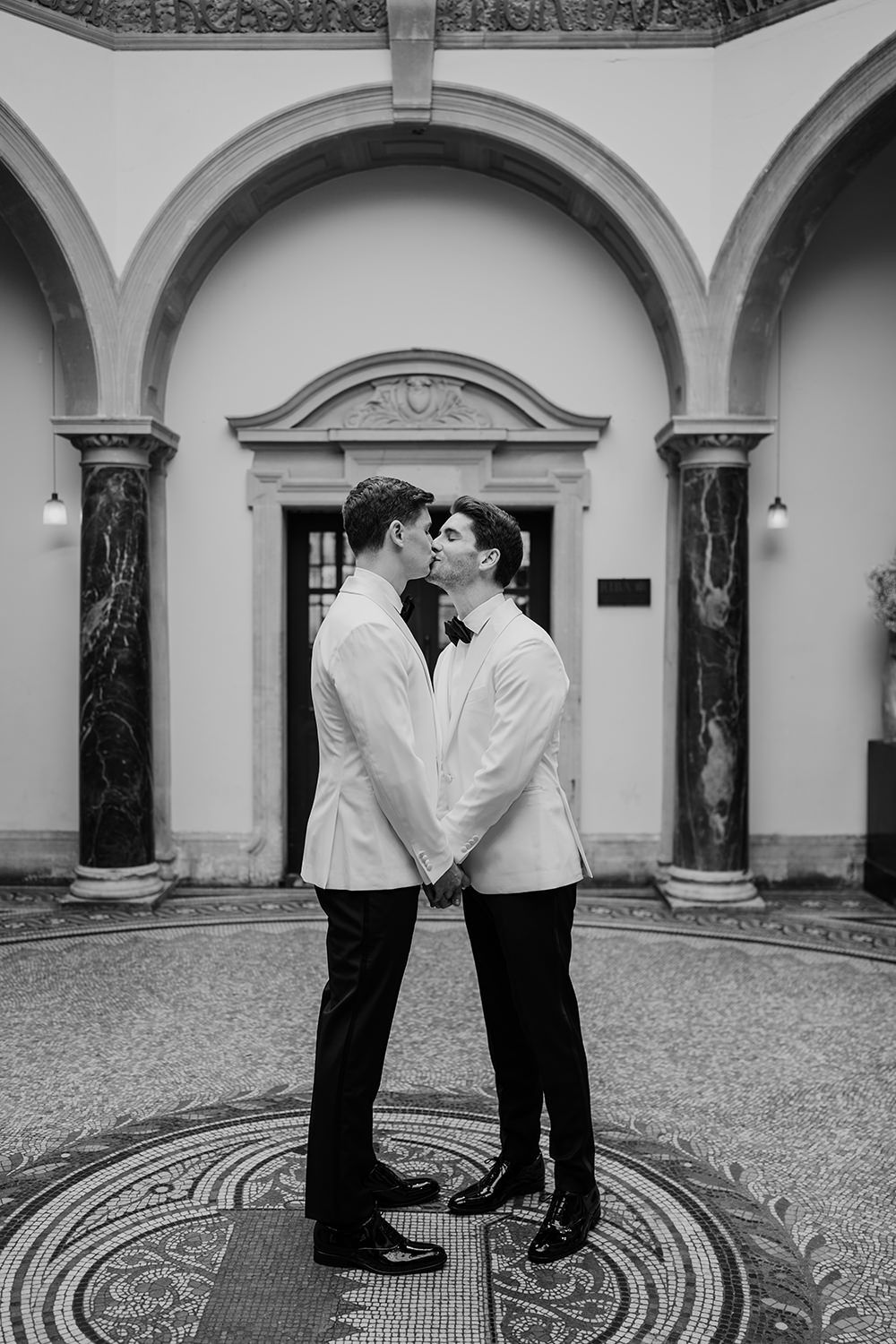 Grooms wearing white and black suits kissing on their wedding day with the architecture of Battersea Arts Centre.