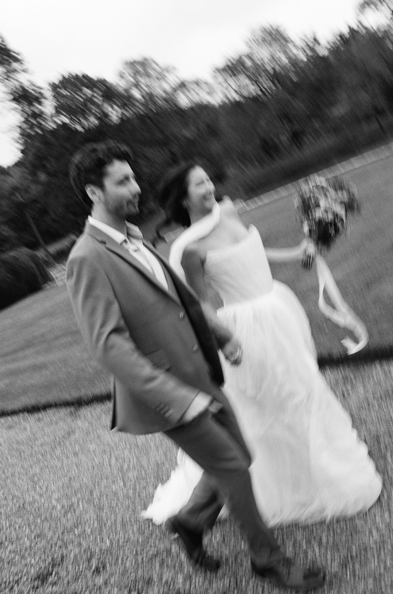 Black and white photo of a joyful bride and groom walking through a field on their wedding day.