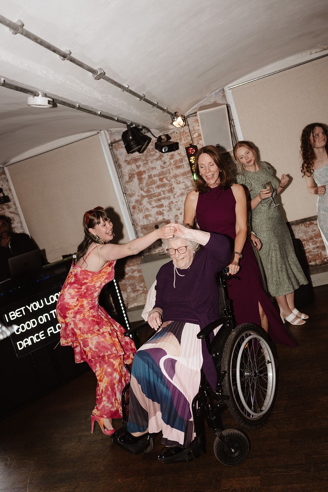 Elderly woman in wheelchair being spun on dance floor at wedding reception by joyful female guests.