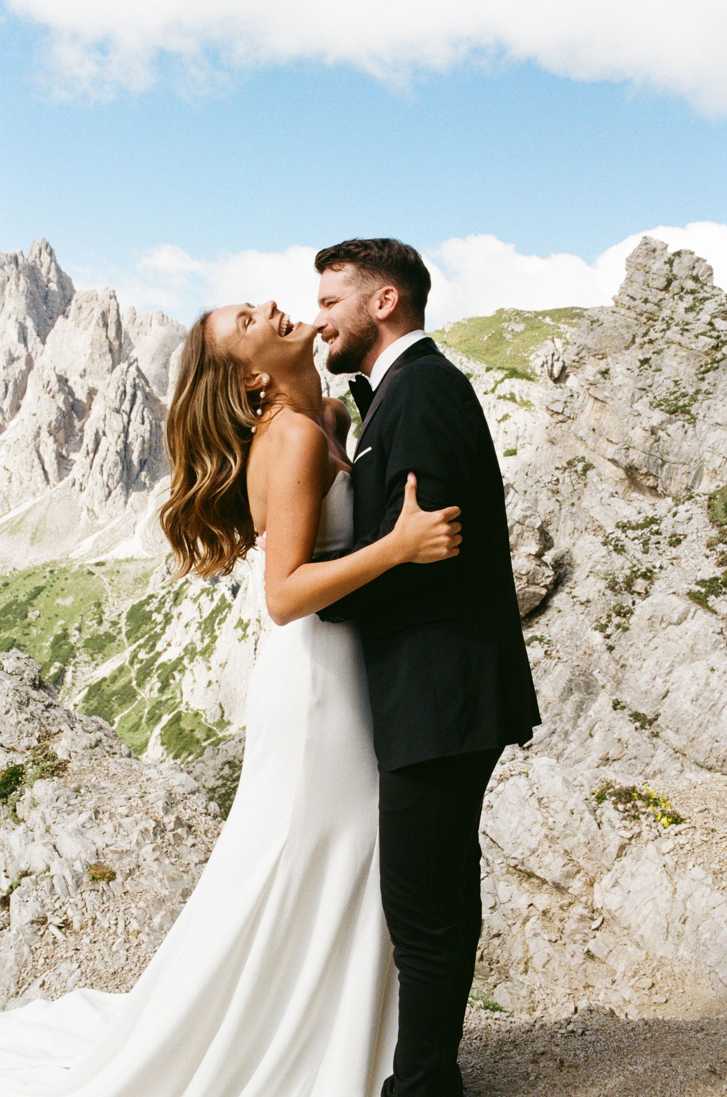 Laughing bride and groom embrace with stunning Dolomite mountain peaks under blue sky as backdrop.
