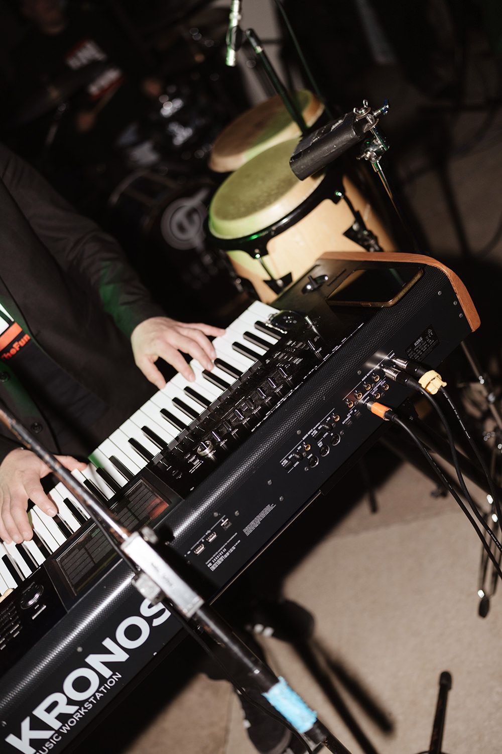 A musician plays a Kronos digital keyboard synth with drums visible in the background during a performance.