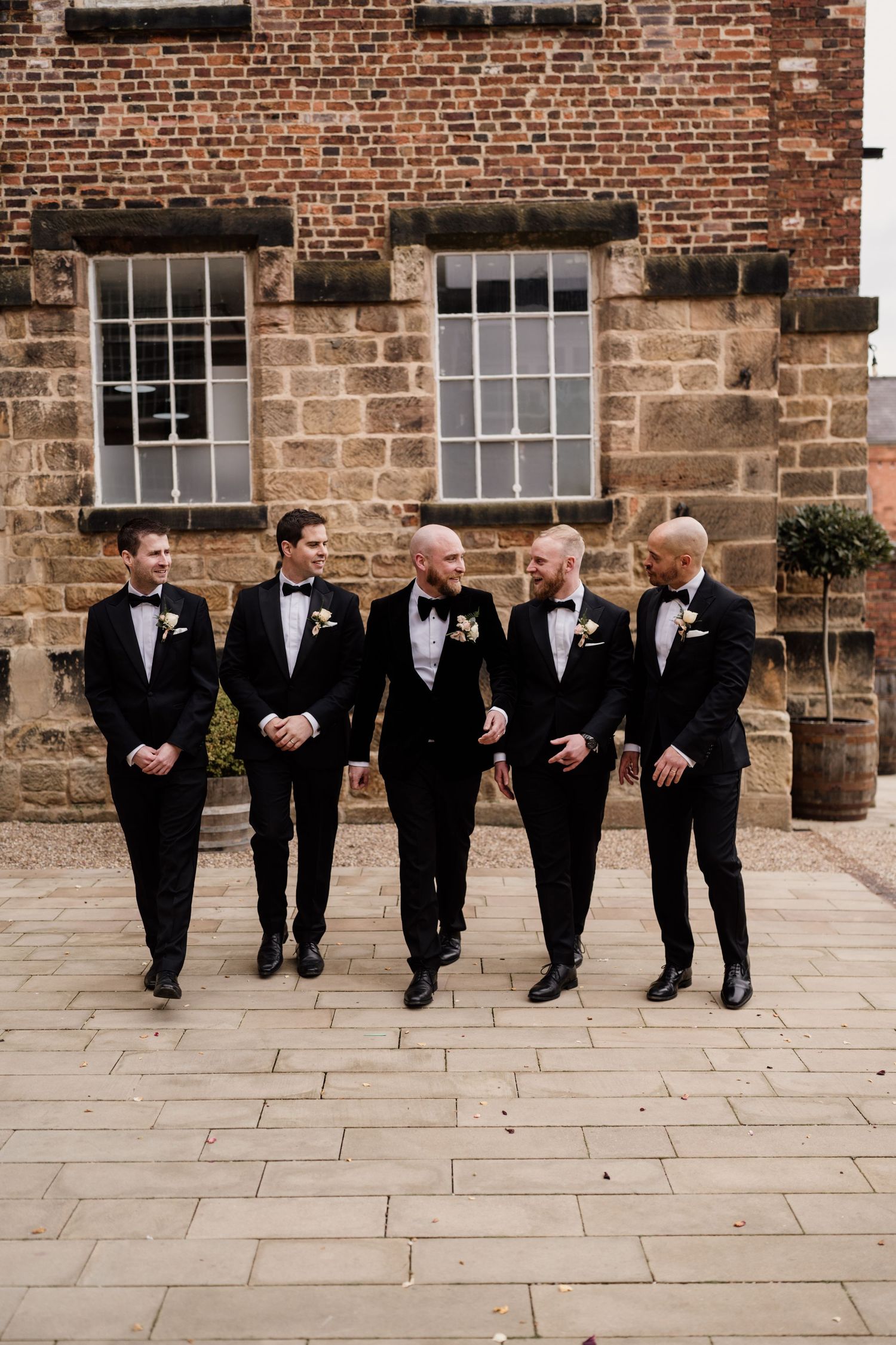 Five groomsmen in black tuxedos pose against a rustic brick wall.