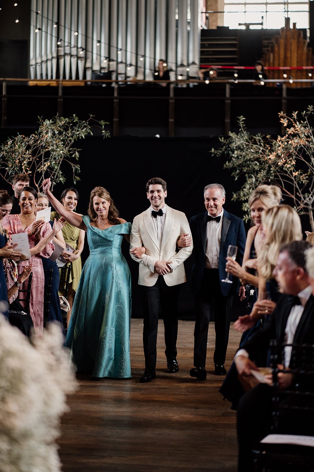 Groom holding his parents hands as he walks down the aisle, holding hands with guests cheering.