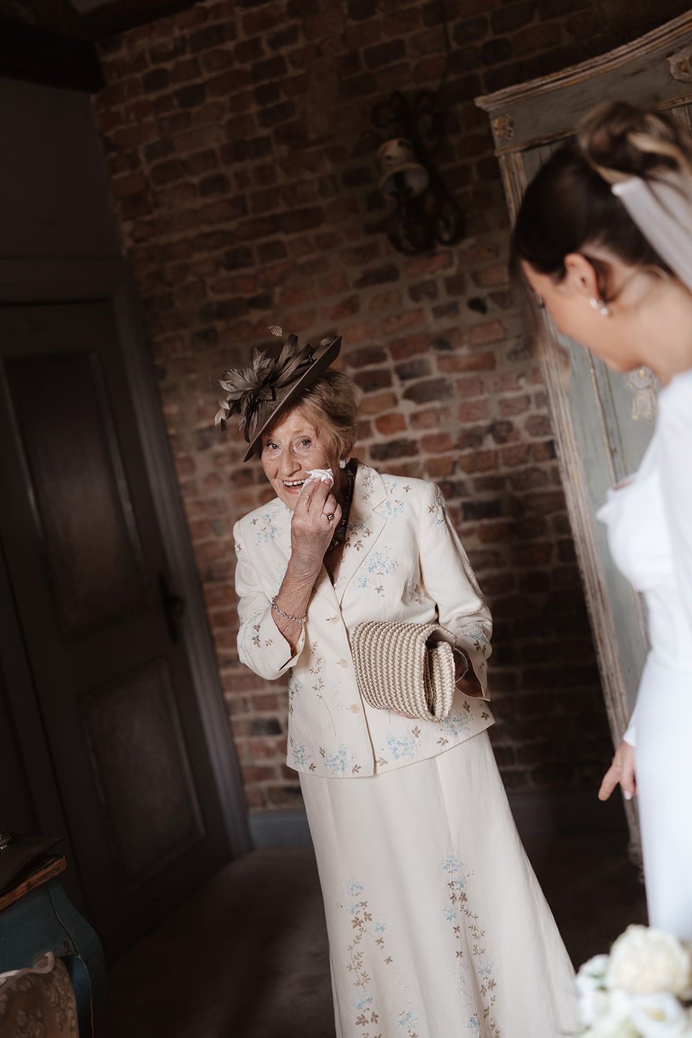 Two people share an emotional moment while wearing formal attire against a rustic brick wall background.