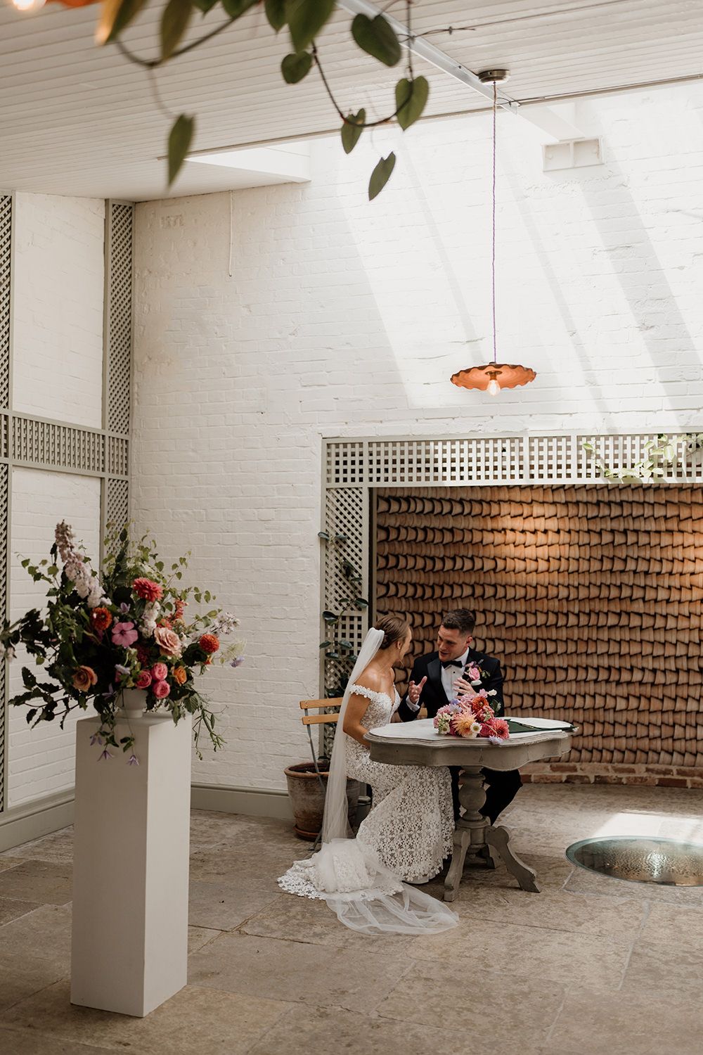 Newlyweds share an intimate moment at a rustic reception table with brick walls and elegant floral arrangements.