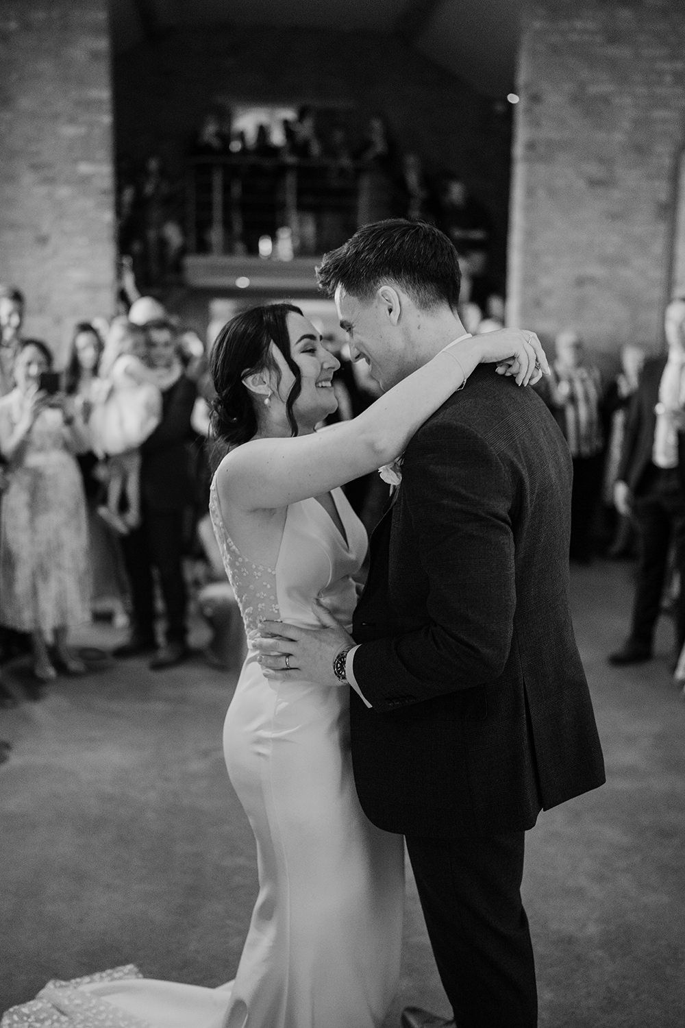 Bride and groom with arms around each other smiling as they do their first dance with friends and family watching.