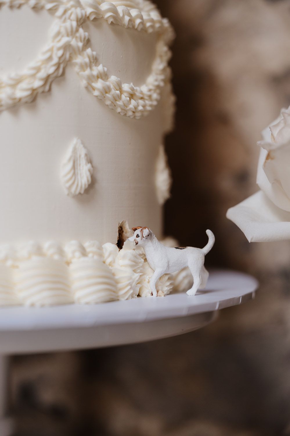 A miniature white dog figurine stands on the edge of an elegant white wedding cake decorated with piped details.