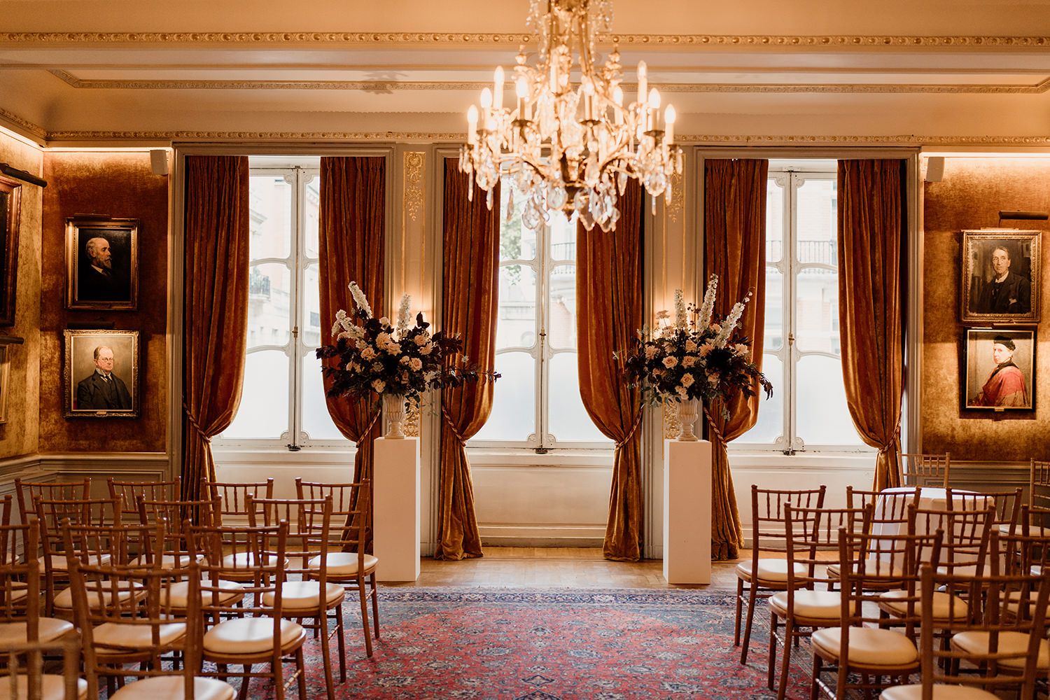 Elegant wedding ceremony setup in grand room with crystal chandelier, wooden chiavari chairs and floral arrangements.