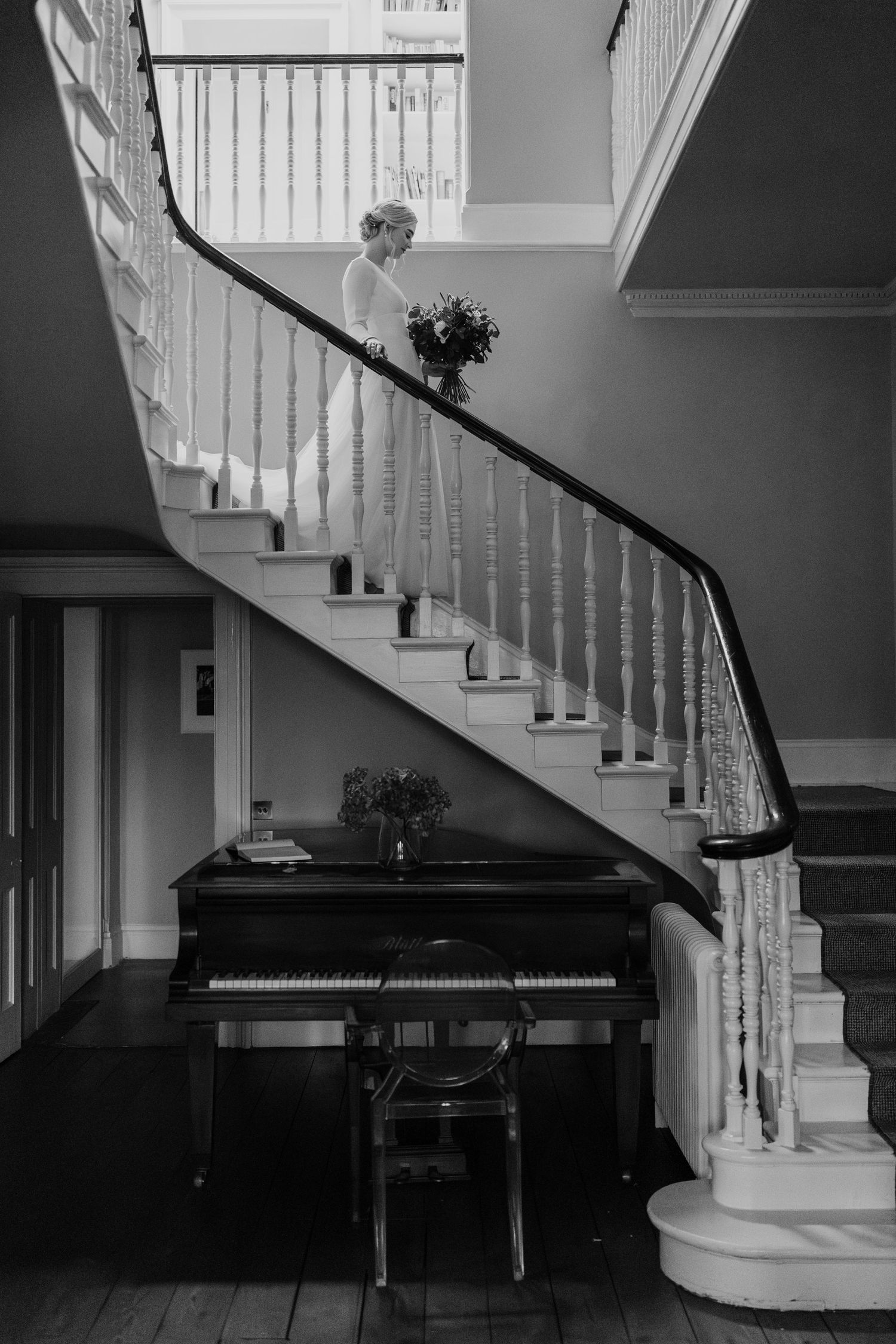 A black grand piano sits beneath an elegant curved white staircase in a sophisticated monochrome interior.