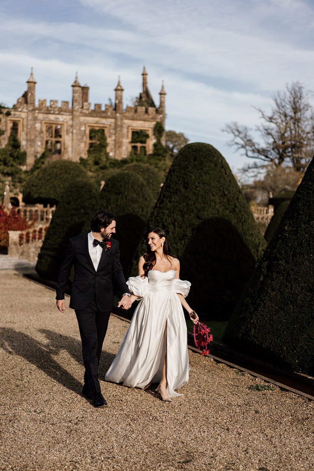 A couple walks along a gravel path beside manicured hedges with a gothic castle-style building in the background.