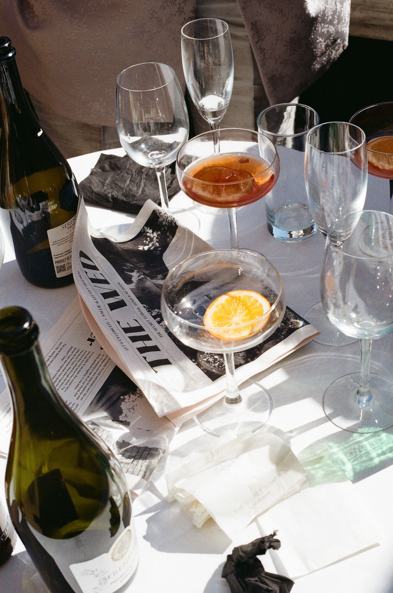 Sunlit wedding reception table with cocktails, wine bottles, and a newspaper centerpiece.