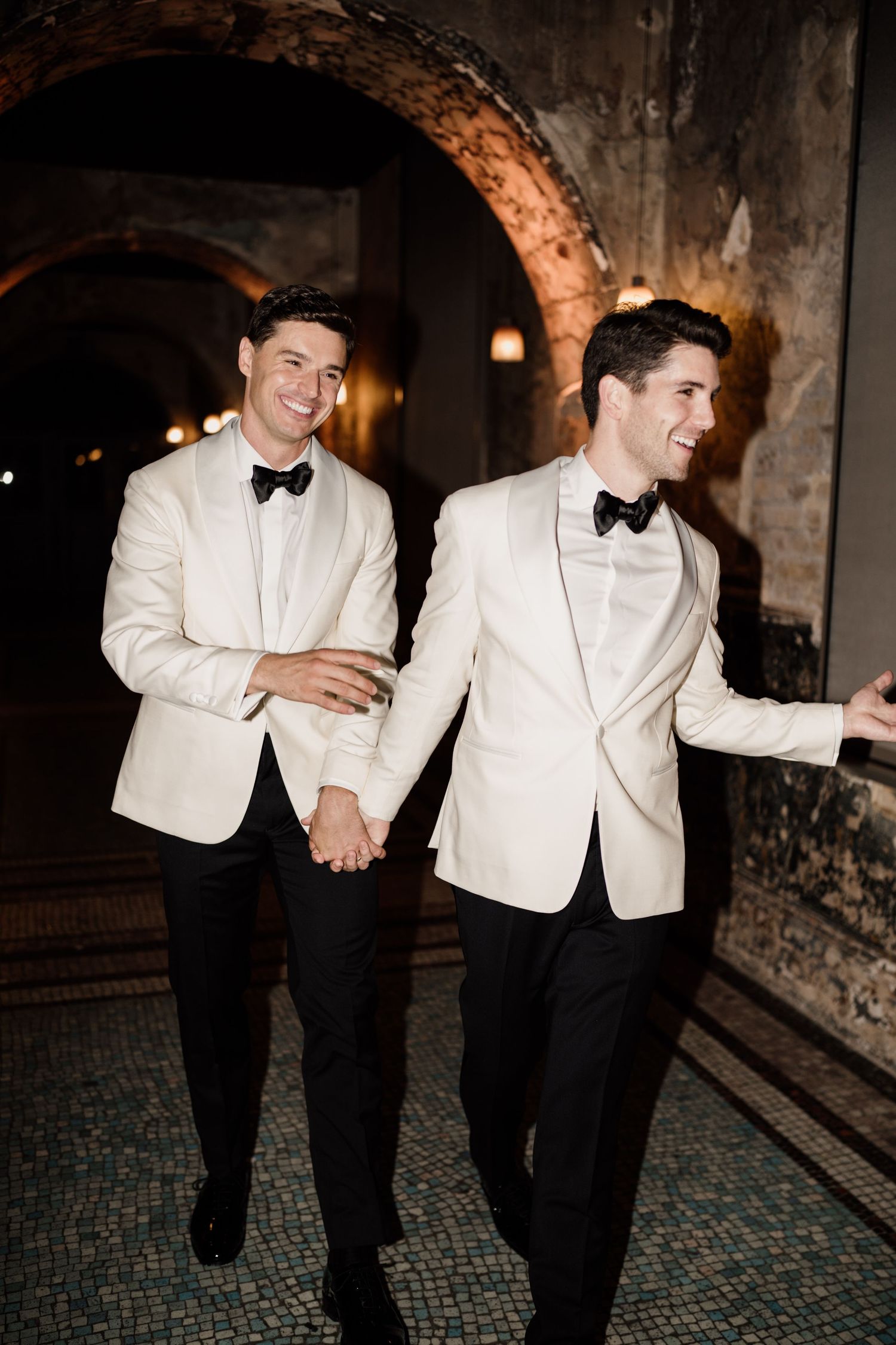 Two grooms in white tuxedo jackets hold hands and smile walking through an arched venue at night.