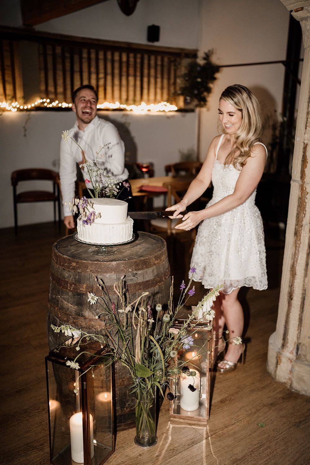Couple cutting wedding cake, the cake is on a wine barrel and the bride is wearing a shorter white dress.