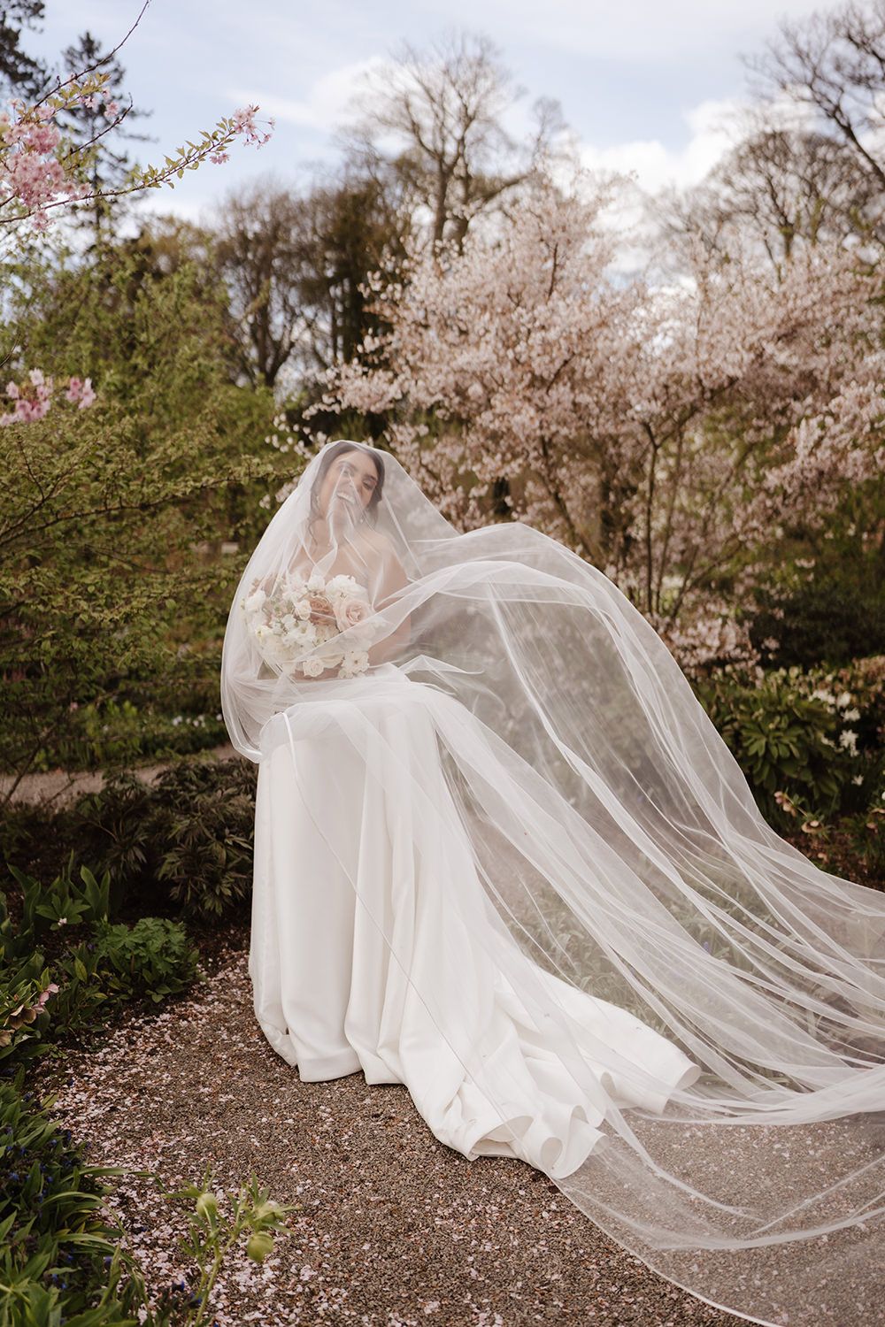 A bride in a flowing white gown and dramatic long veil poses among cherry blossoms in a spring garden setting.