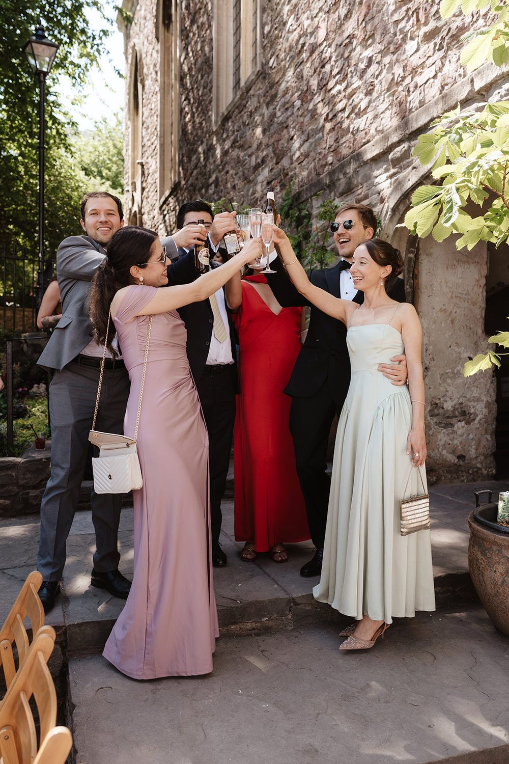 A celebratory group gathers outdoors against a stone wall, raising glasses in a festive toast.