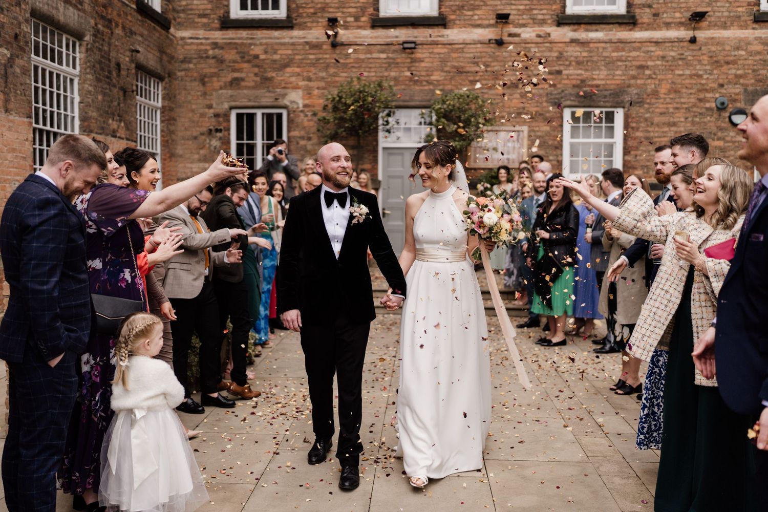 Wedding party celebrating with confetti toss outside historic brick venue.