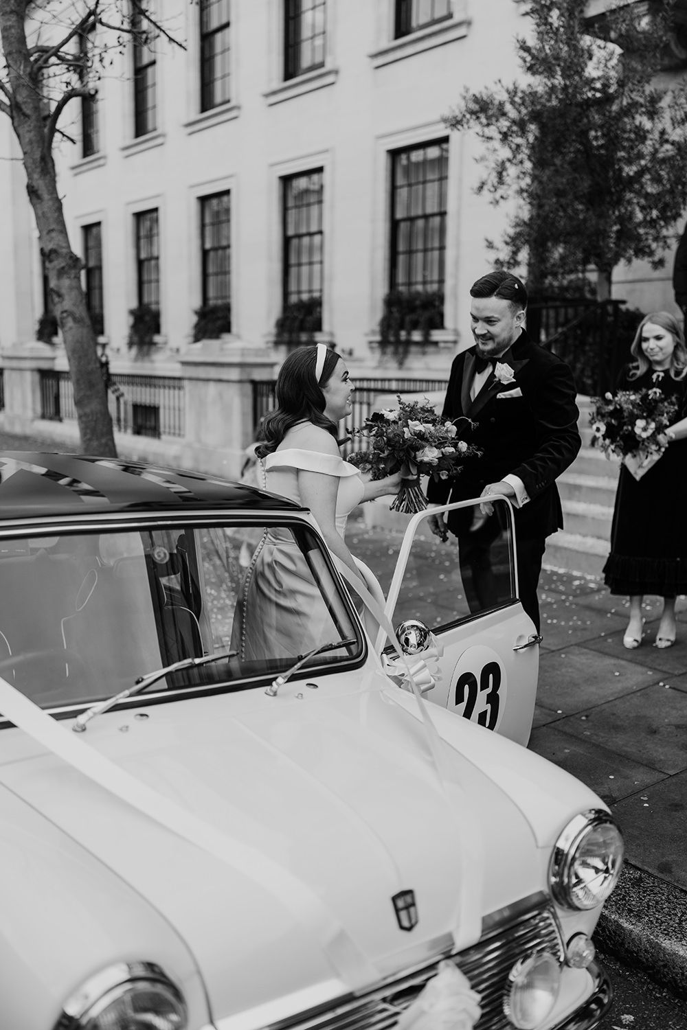 A vintage car numbered 23 is parked on a street beside a classic building in black and white photography.