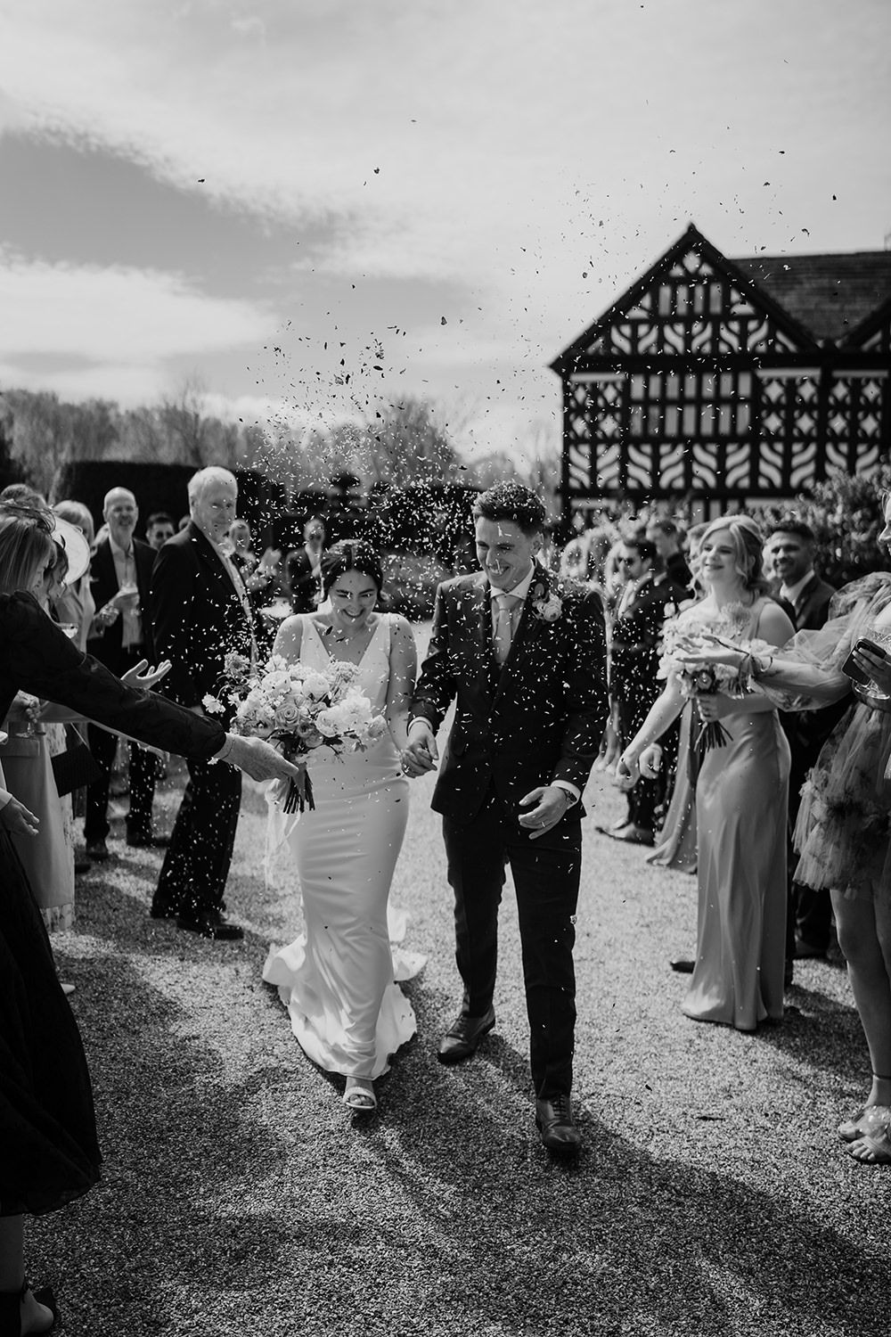 Black and white image of bride and groom walking amongst confetti with Holford Hall behind them as guests cheer.