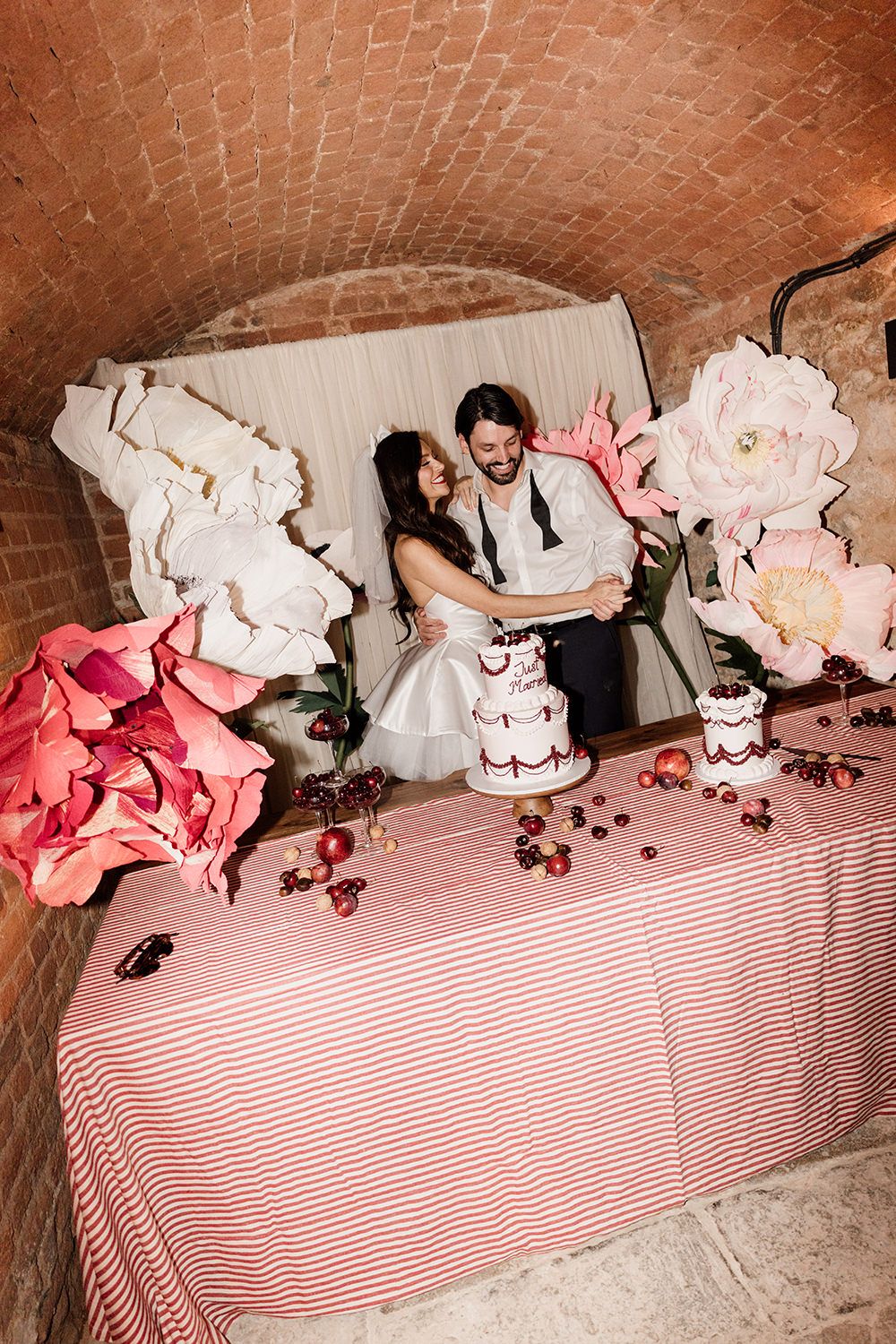 Romantic wedding couple sits at table with pink and white paper flowers and striped tablecloth in rustic brick cellar.