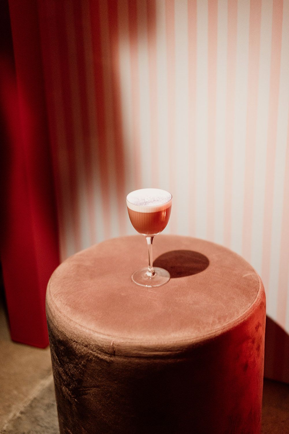 A wine glass sits atop a rich brown velvet ottoman or cushion in a warm-toned room with striped wallpaper.