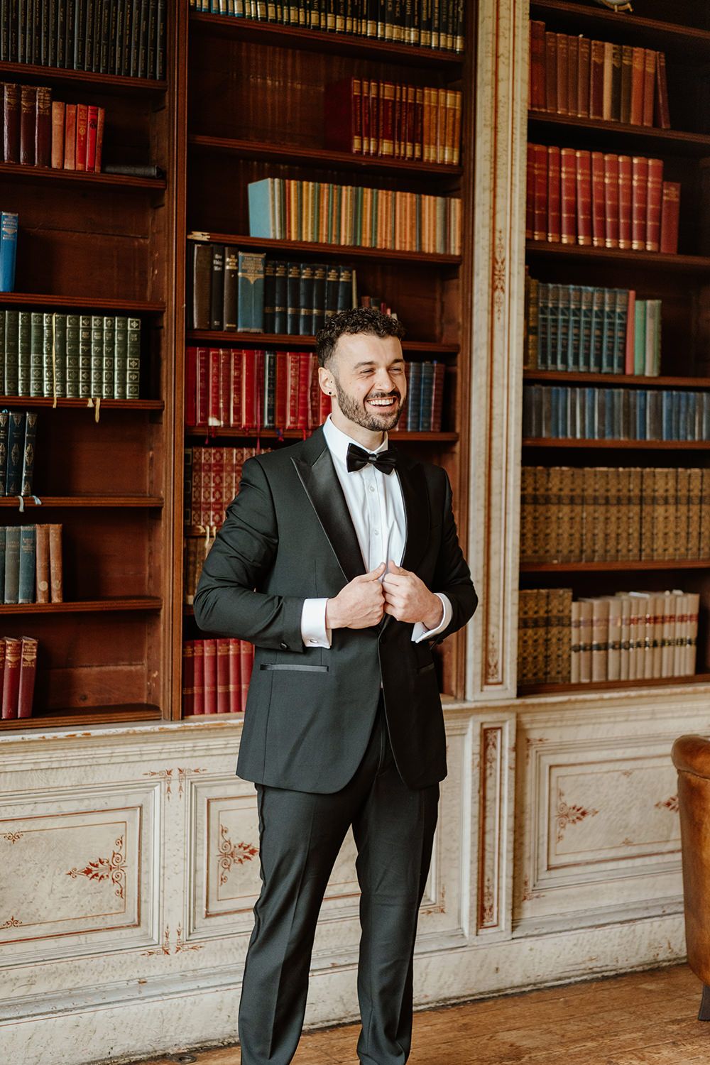 Groom adjusting his suit while smiling in the library at Rise Hall.
