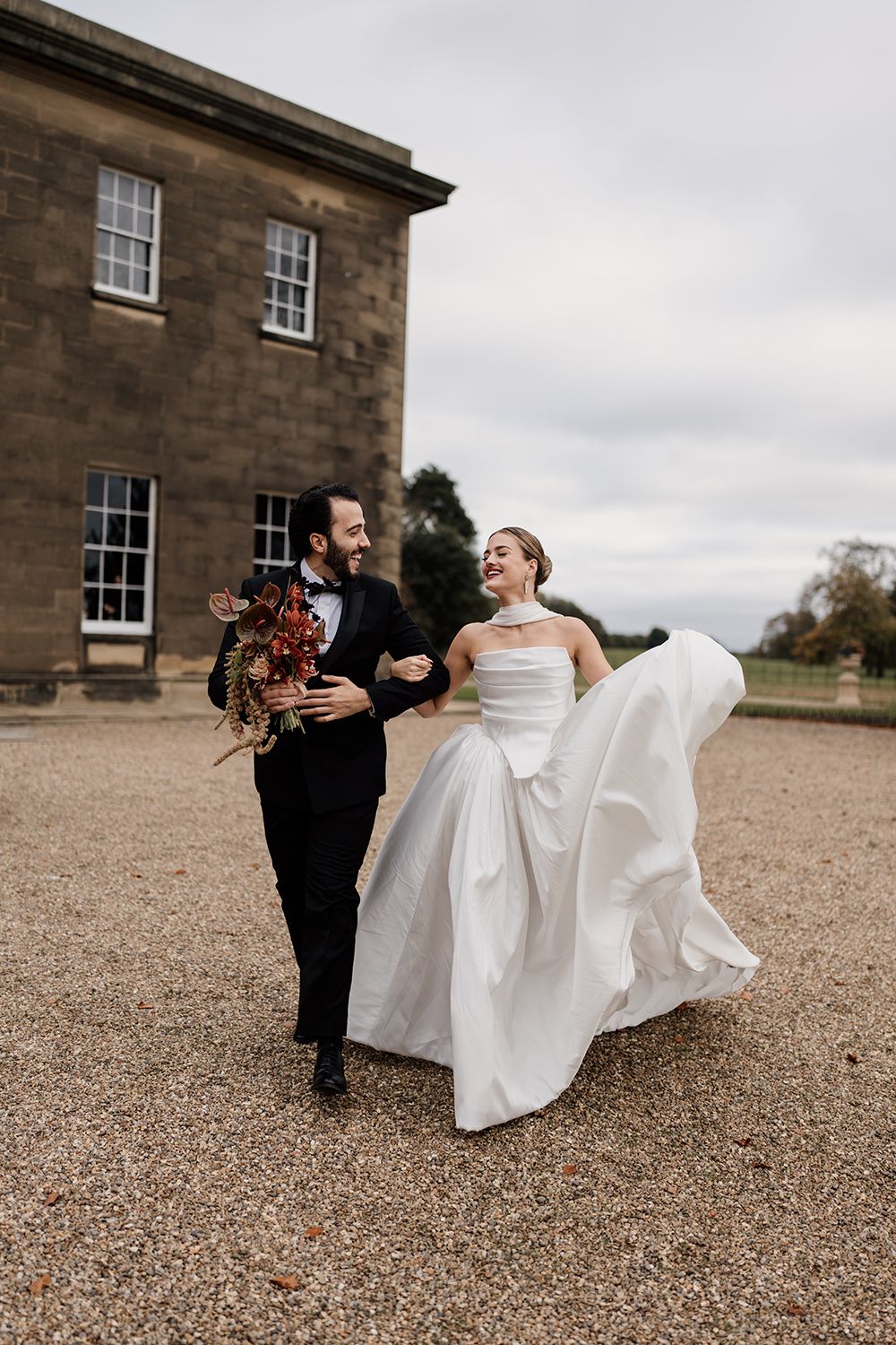 Bride and groom running in the grounds of Rise Hall, holding autumnal flowers while laughing and smiling.