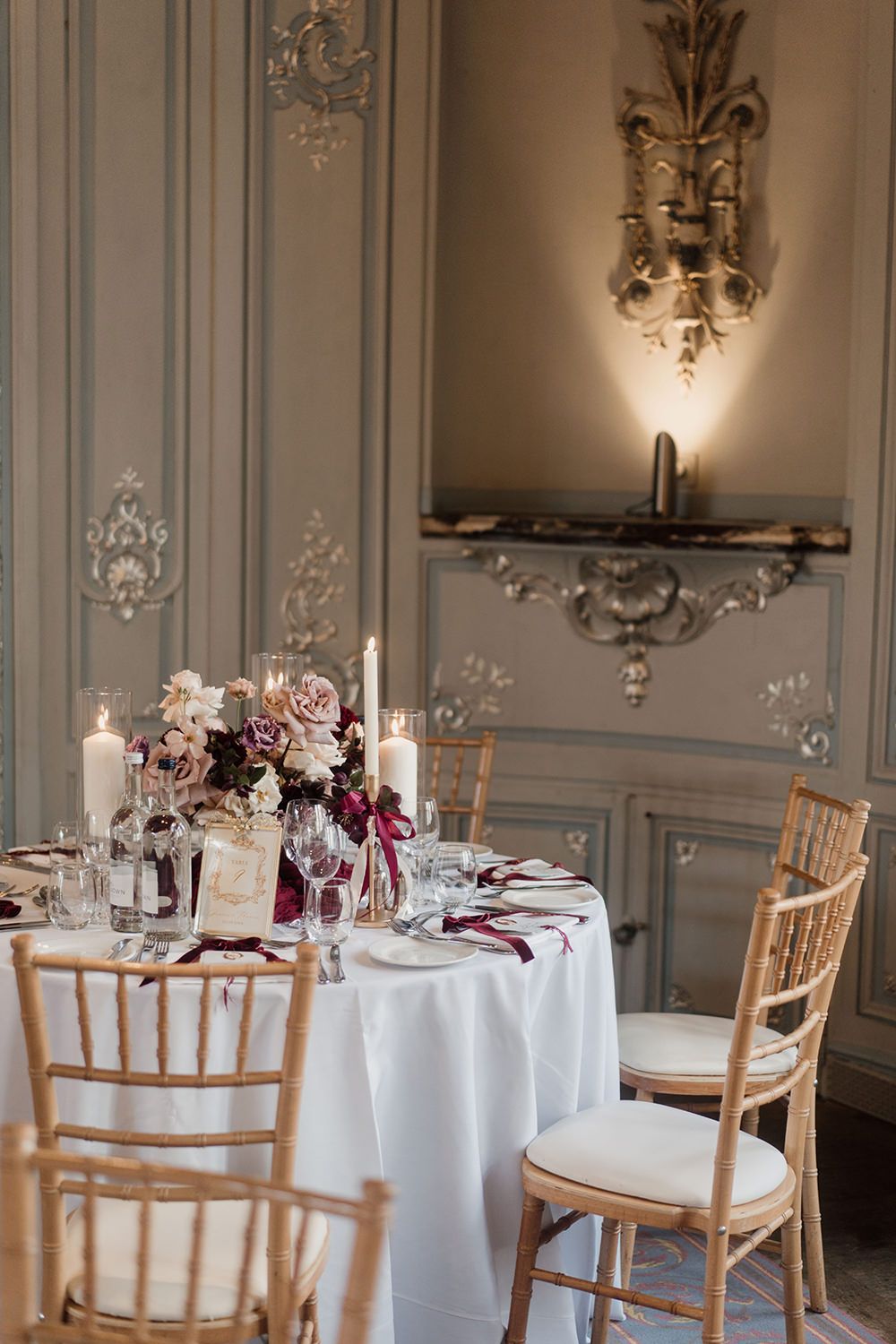 Elegant wedding table setting with burgundy floral centerpiece and chiavari chairs in luxurious French-style room.