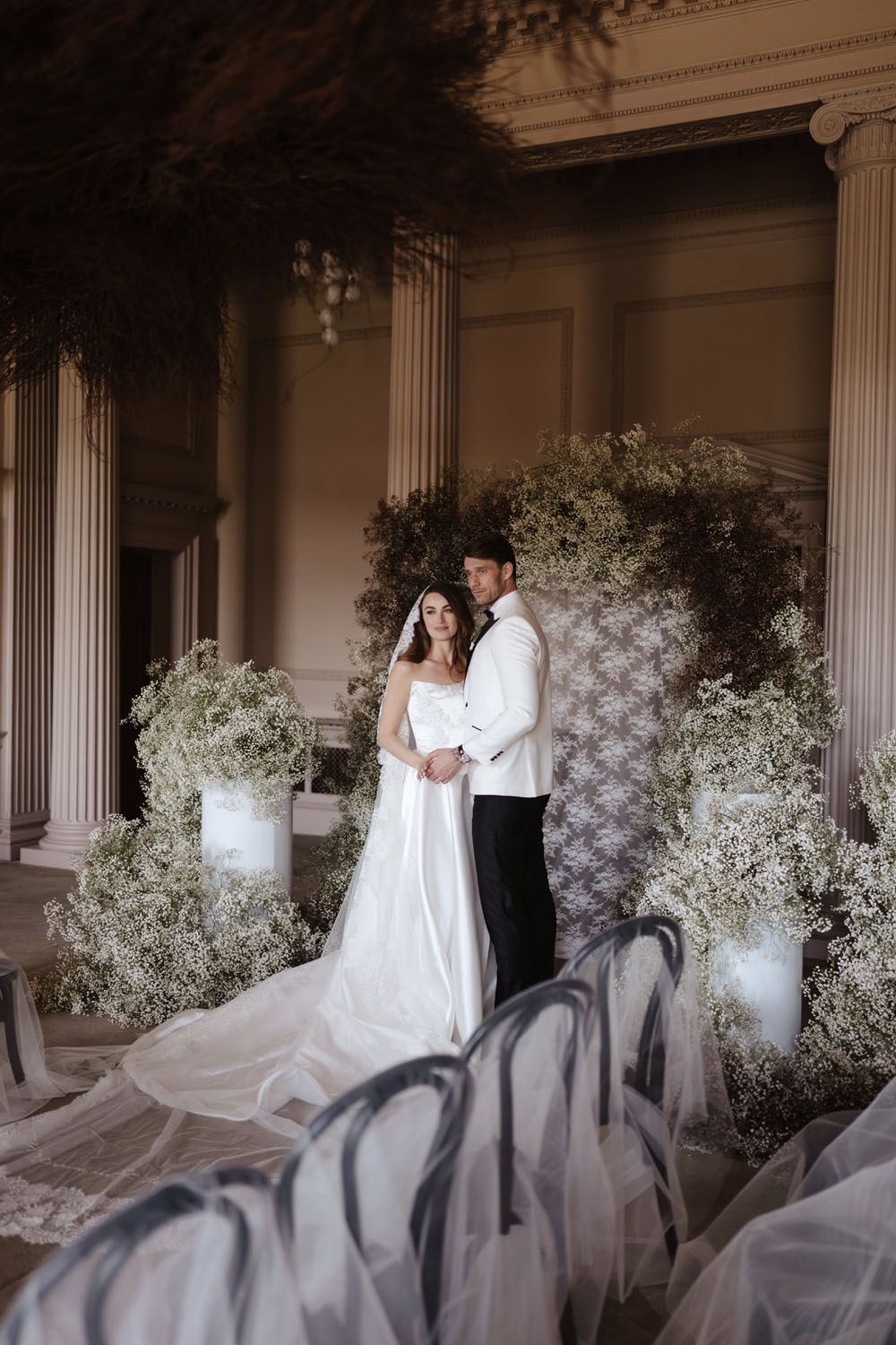 Bride and groom embracing at elegant wedding ceremony with lush white floral arch and classical columns.