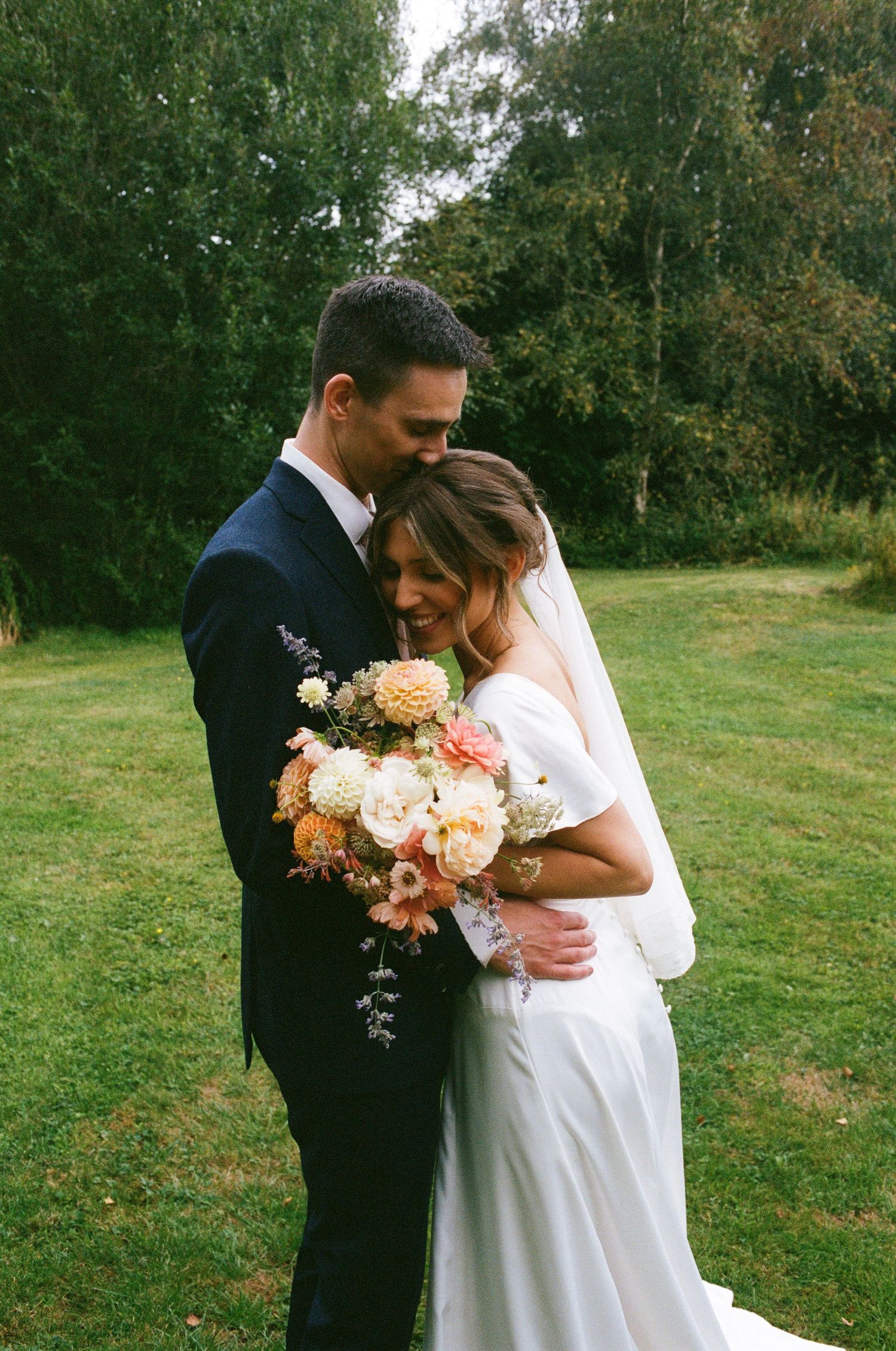 A bride and groom embrace outdoors on green grass, she holds a peach and white floral bouquet, wearing a white gown and veil.